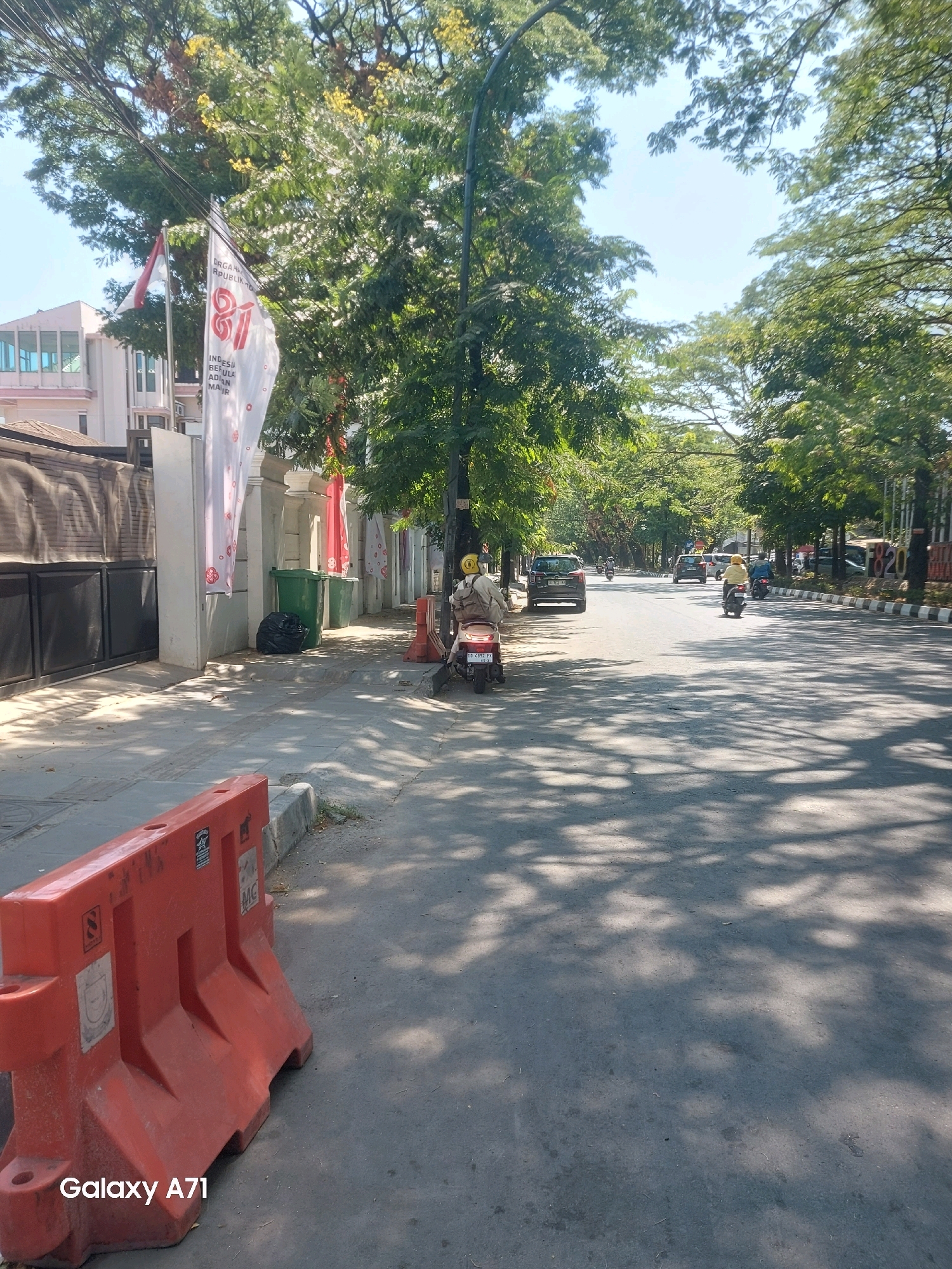 The image depicts a street scene in Makassar, Indonesia, on a sunny day. The foreground features a bright orange traffic barrier, a common sight on Indonesian streets. The sidewalk next to the street is lined with trees, casting dappled shadows on the pavement. On the sidewalk, there are two green trash bins, one of which has a black garbage bag next to it.
Further down the street, several motorcycles and cars are visible. One motorcycle with a rider wearing a yellow helmet is parked on the side of the road. Another motorcycle with a rider also in yellow is moving along the street. A dark-colored car is parked on the left side of the road. Further in the distance, more vehicles are on the road, and the street is lined with trees and buildings.
Two flags are prominently displayed near the sidewalk. One is the Indonesian flag, red and white. The other is a white flag with a large "81" graphic and text that appears to be in Indonesian, possibly commemorating an event or anniversary. The building in the background on the left appears to be a multi-story structure with modern architecture.
The overall impression is of a typical urban street in Makassar, with everyday activity and elements of local culture and events represented by the flags.