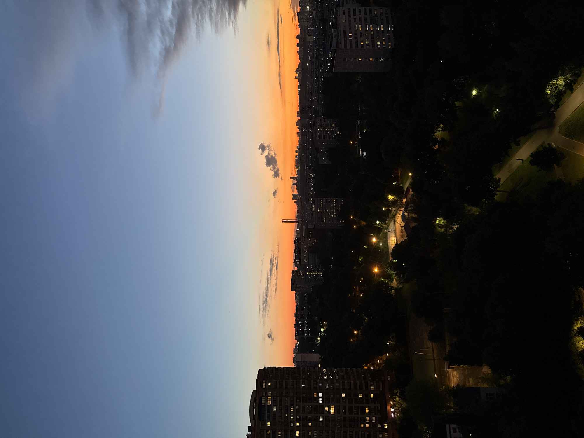 This is a long-shot, vertical, full-frame, eye-level, outdoor, natural-light photograph taken at dusk, looking east over a dense urban landscape in Moscow, Russia. The sky displays a gradient from a pale blue at the top to a vibrant orange and red near the horizon, indicating the sunset. Scattered clouds, some tinged with orange, are visible in the sky. In the mid-ground, numerous apartment buildings of varying heights and architectural styles are illuminated by interior lights. A prominent, multi-story apartment building with many windows is situated on the left side of the frame. The foreground is dominated by the dark silhouettes of trees and parkland, with streetlights casting pools of yellow light on pathways. There are no discernible people or specific interactions depicted in the scene. The overall impression is of a sprawling residential area of Moscow at the transition from day to night.