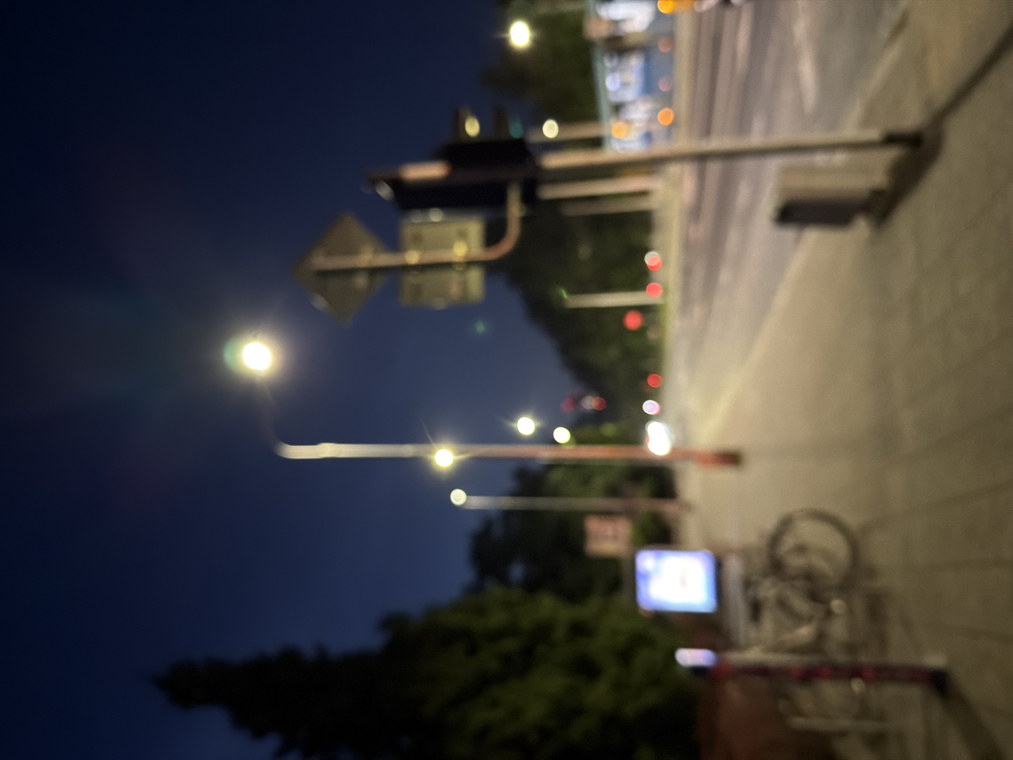 An out-of-focus, nighttime, street-level shot captures a sidewalk and an adjacent street in an urban environment. Several streetlights illuminate the scene, casting a soft glow. A traffic light is visible, with its signal lights appearing as blurred orbs of color. On the sidewalk, a bicycle is parked next to a glowing billboard. Trees line the background, silhouetted against the dark sky. The overall impression is one of a quiet, dimly lit street at night.