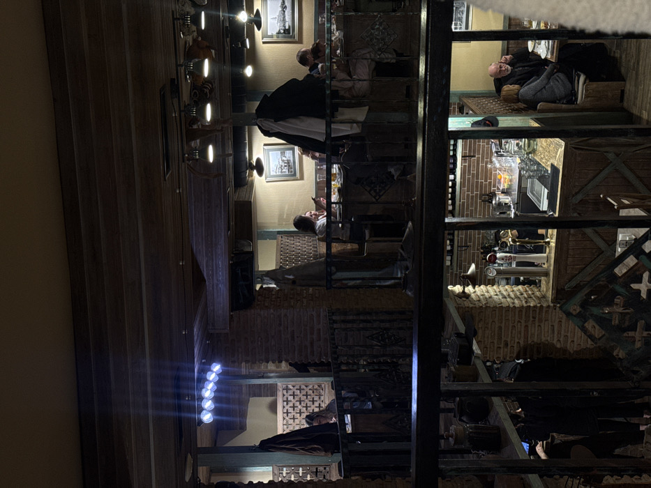 The image depicts the interior of a multi-level restaurant or bar in Tbilisi, Georgia, viewed from an elevated position. The establishment features a rustic design characterized by dark wooden ceilings, beams, and railings, complemented by exposed brick pillars and walls.
In the mid-ground, a mezzanine level is visible with multiple patrons seated at tables. On the left, a woman is seated at a table within a booth-like structure, partially obscured by a dark coat hanging from a partition. To her right, a prominent horizontal fixture with seven bright circular white lights is mounted on a wooden beam. On the right side of this level, two women are seated at a table, one of whom is actively looking at a mobile phone. Other patrons are partially visible further back. Coats are draped over chairs or hanging on hooks throughout this area. Framed pictures hang on the light-colored walls.
Below this mezzanine, a lower bar area and additional seating are visible. The bar features stainless steel elements, at least three beer taps, various bottles, glasses, and a bowl of citrus fruits. Adjacent to the bar, on the far right of the image, an older man with a beard is seated at a table, holding a phone to his ear. Across from him, an older woman is also seated, engaging in conversation.
The overall lighting is dim and ambient, provided by various overhead fixtures, creating a warm atmosphere, with the exception of the aforementioned bright white circular lights. Ornate dark metal railings divide the different levels and sections.