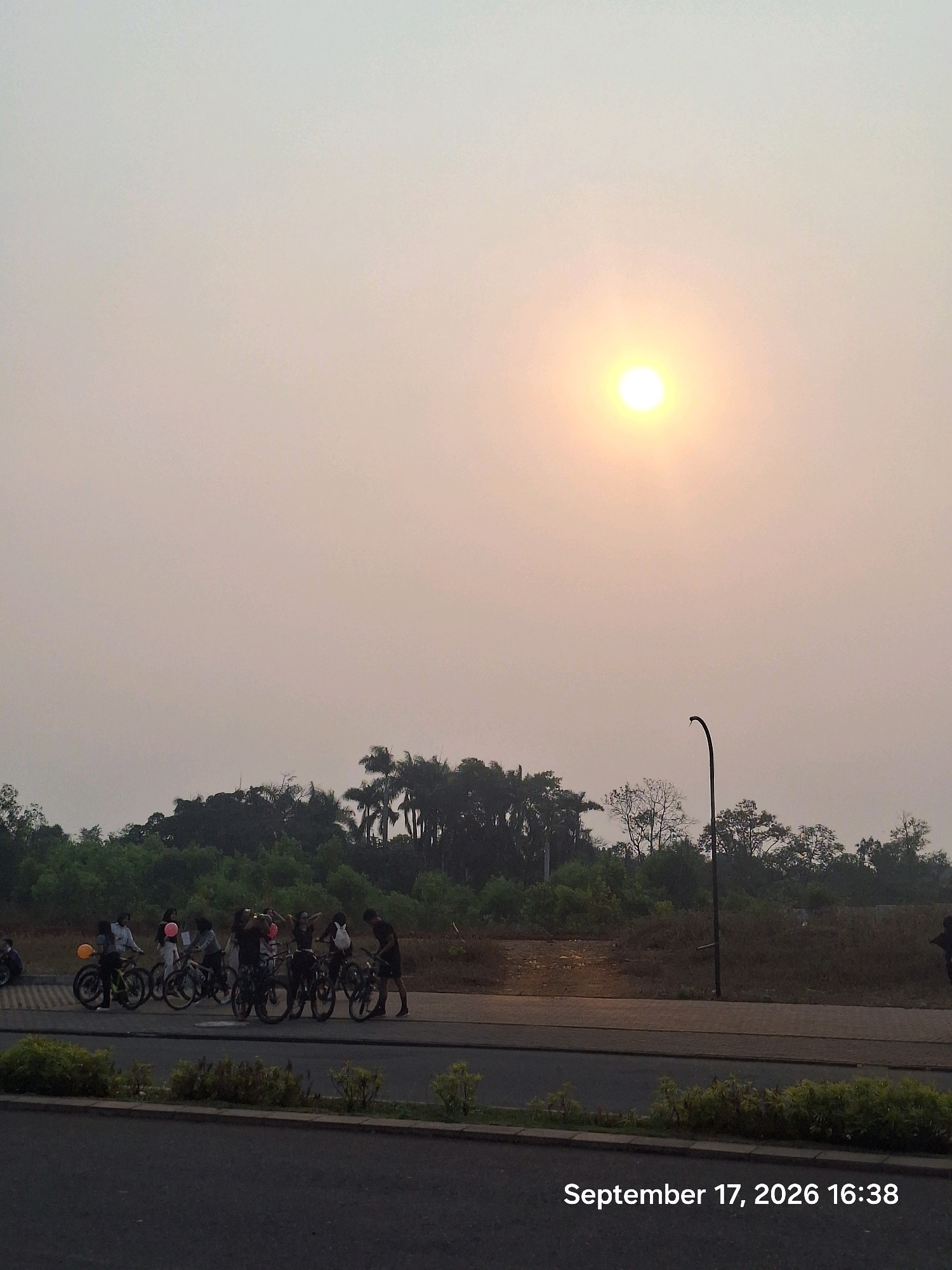 An outdoor, eye-level shot captures a hazy sunset over a park-like area in Depok, Indonesia. The sun, a bright yellow orb, dominates the upper right quadrant of the sky, casting a soft, diffused light. Below the sky, a dense line of trees forms the horizon, with palm trees visible among them. In the middle ground, a group of approximately eight individuals are gathered with bicycles. Some appear to be standing and talking, while others are positioned near their bikes. One person on the far left is partially obscured by a balloon. The group is positioned on a paved pathway that runs horizontally across the frame. In the foreground, a dark asphalt road is visible, with a strip of green vegetation and a concrete barrier separating it from the pathway. The timestamp "September 17, 2026 16:38" is overlaid on the lower right corner of the image.