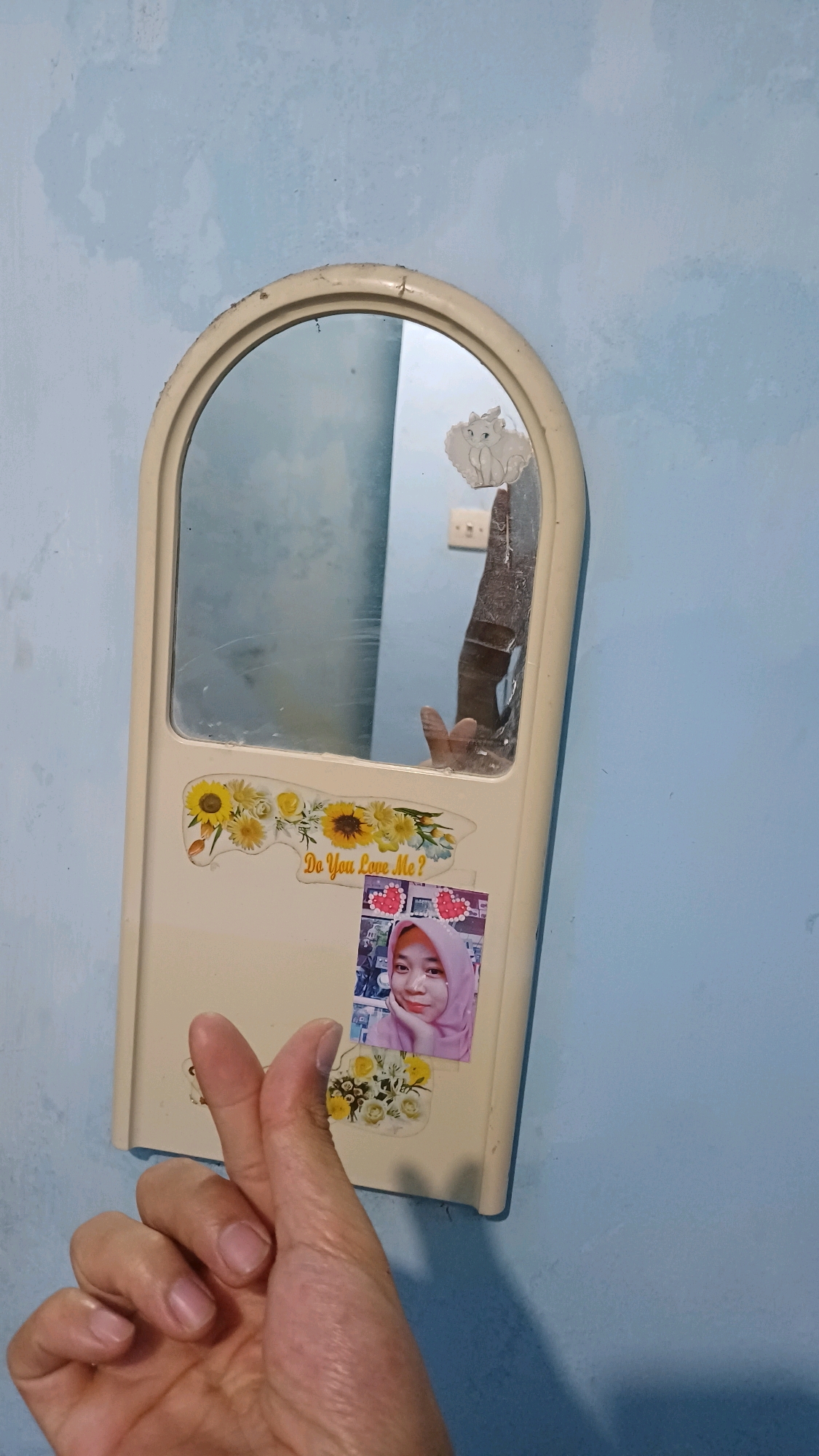 A person's left hand is in the foreground, with the thumb and index finger forming a heart shape. The hand is positioned in front of a cream-colored, arched mirror hanging on a light blue wall. The mirror reflects a portion of the room and a person's arm and hand. On the mirror, near the bottom left, there is a sticker with sunflowers and the text "Do You Love Me?". Below this sticker, there is a small photograph of a woman wearing a pink hijab. Another sticker depicting a white cat is visible on the top right of the mirror. The wall has a textured, slightly faded appearance. The scene is indoors, likely in a domestic setting in Kowangan, Indonesia.