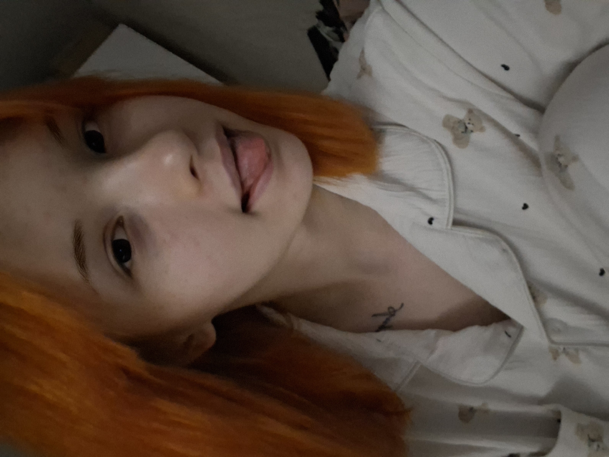 The image is a close-up, vertical selfie featuring a person with short, bright orange hair. The subject is looking directly at the camera with a playful expression, sticking out their tongue slightly. They appear to be lying down, likely in a relaxed indoor setting such as a bed.
The person is wearing light-colored, long-sleeved pajamas or a button-up shirt. The fabric of the garment is patterned with small, light brown bear figures and tiny black dots. A small, dark tattoo, appearing as a stylized letter or symbol, is visible on their left collarbone or upper chest area. The background is dark and out of focus, providing no specific location cues beyond indicating an indoor environment. The overall mood of the image is informal, relaxed, and lighthearted. The location context for this scene is Chaikovsky, Russia.