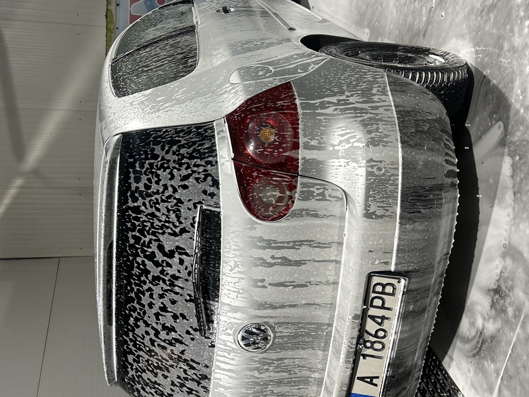 A silver Volkswagen Golf hatchback, identified by the prominent Volkswagen emblem on its tailgate and its characteristic taillights, is actively being washed inside a car wash bay. The entire vehicle is covered in a thick layer of white soap foam, which is visibly dripping down all surfaces, including the rear window, body panels, and circular taillights. The Bulgarian license plate, "A 1864 PB," is clearly displayed on the rear bumper, with the 'A' indicating the city of Burgas. The car features a rear spoiler and a rear window wiper. Small circular parking sensors are visible on the rear bumper. The rear wheel, encased in a black multi-spoke rim, is also covered in foam, resting on a wet, foamy concrete floor. The background consists of light-colored, possibly corrugated metal, walls typical of an indoor car wash facility. No individuals are present in the frame.