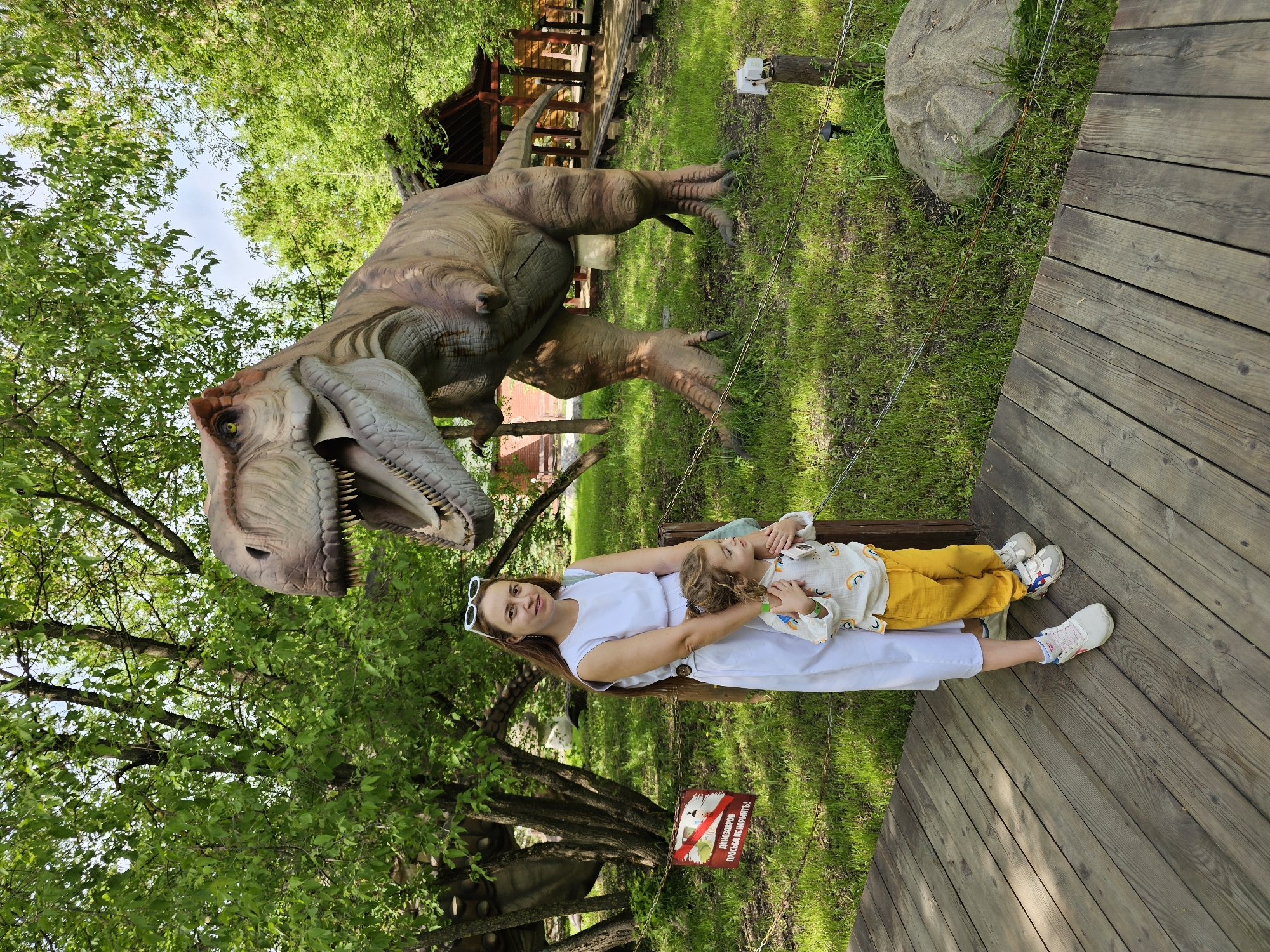 A photograph taken in the city of Khimki, Russia, depicts a woman lying on a wooden platform with a child in her arms. The woman is wearing a white dress and white sneakers. The child is wearing a white shirt with rainbows, yellow pants, and white sneakers. Behind them stands a large model of a Tyrannosaurus Rex dinosaur. The surrounding environment includes trees, grass, and a sign with Russian text and a dinosaur icon. A wooden structure is visible in the background.