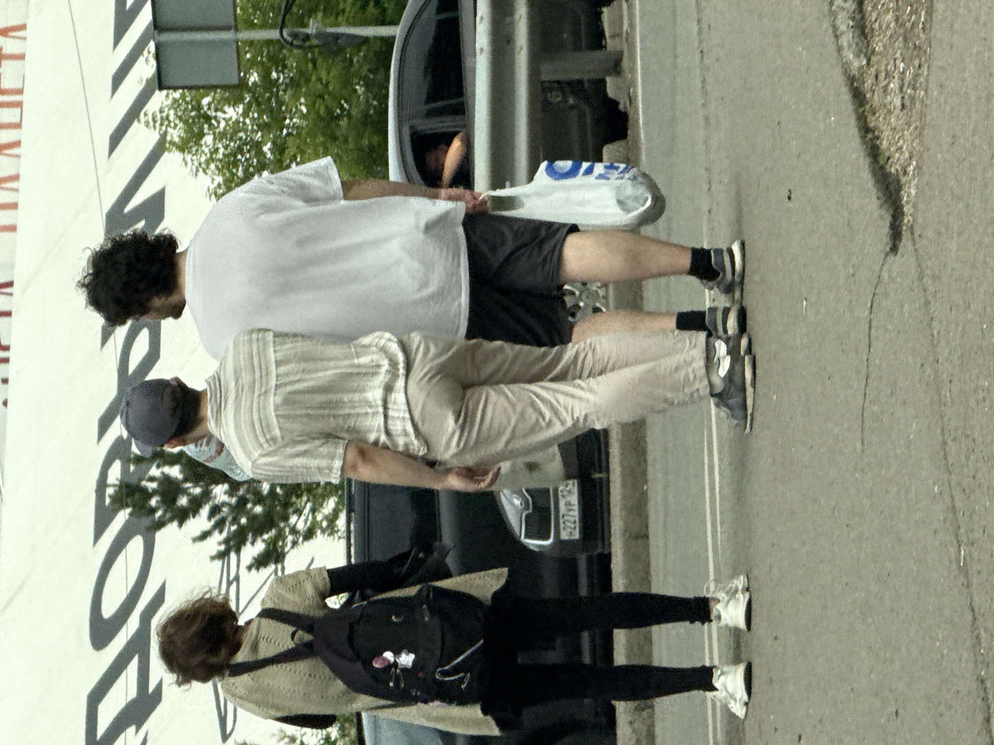 The image depicts three individuals standing near a road in Krasnoyarsk, Russia. The foreground is dominated by the asphalt surface of the road. In the middle ground, three people are visible from the waist down, facing away from the camera. To the left, a woman wearing black leggings and white sneakers stands with a black backpack on her back. In the center, a person in light-colored pants and grey and white sneakers is seen. To the right, another person in black shorts and black socks with grey sneakers stands, holding a white plastic bag with blue lettering. Behind the individuals, a dark-colored car is parked on the road, and a metal guardrail runs along the side. In the background, a large white banner with dark lettering is displayed, and lush green trees are visible. The overall scene suggests a public outdoor setting in Krasnoyarsk.