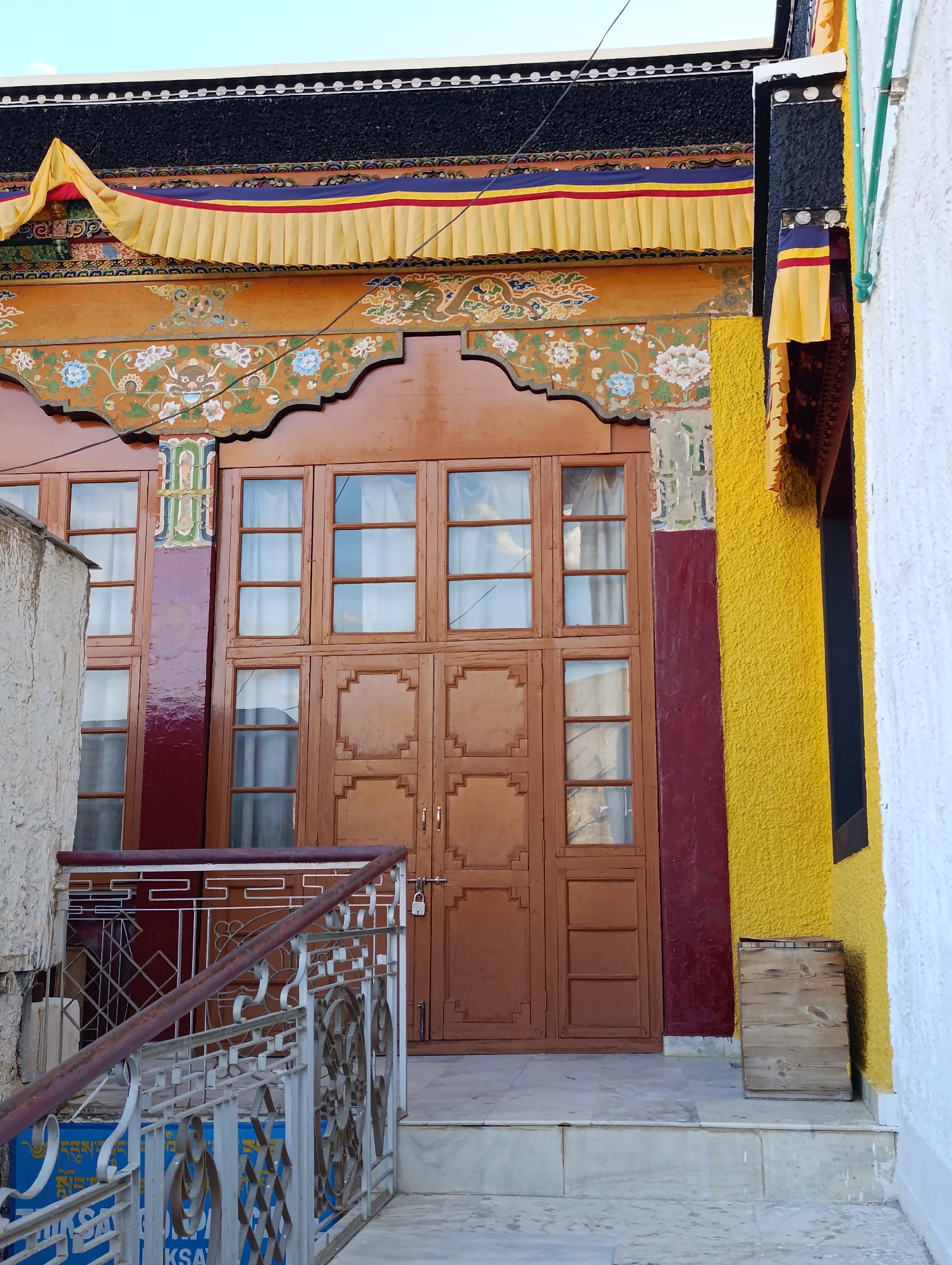 The image shows a close-up view of the entrance to a building with traditional Tibetan architectural elements. The building features a large wooden door with multiple panels and windows, decorated with ornate carvings and colorful paintings. Above the door, a series of decorative elements include pleated fabric banners in yellow, red, and blue, as well as painted motifs of flowers and mythical creatures. To the right of the entrance, a section of the building is painted bright yellow, contrasting with a white wall on the far right. A wooden crate is placed on the ground near the yellow wall. A metal railing with intricate designs is visible in the foreground, leading up to the entrance. The overall scene suggests a cultural or religious site, likely in a Tibetan Buddhist region.