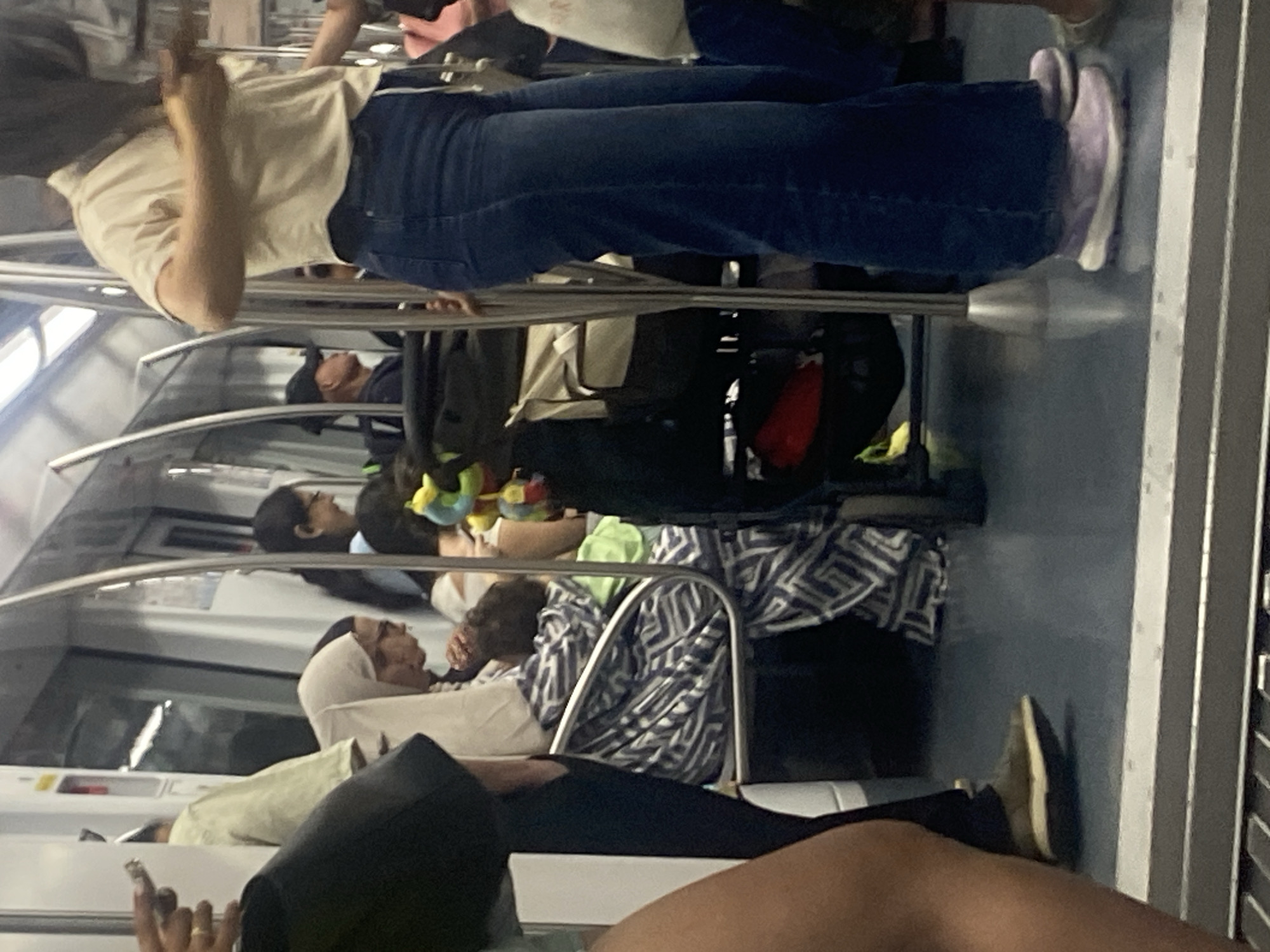 An interior view of a subway train in l'Hospitalet de Llobregat, Spain, shows several passengers seated and standing. A person in the foreground, wearing a cream-colored shirt and blue jeans, stands facing away from the camera, holding onto a vertical pole. To their left, a woman wearing a white hijab is seated, holding a baby wrapped in a blue and white patterned blanket. Another person, wearing glasses, is seated next to the woman with the baby. In the background, other passengers are visible, including a person wearing a black baseball cap. Bags and luggage are stowed near the seats. The floor of the train car is a dark, reflective blue, and the walls are a light color with visible metal poles for support.