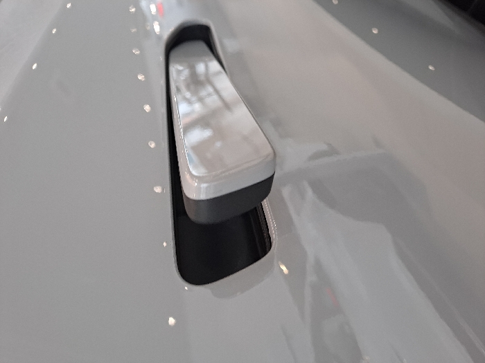 A close-up depicts a flush door handle, possibly of a car. The handle's top is reflective silver, contrasting with a darker gray underside. It's embedded within a light gray surface, presumably a car door or panel, creating a smooth, aerodynamic appearance. Light reflections accentuate the curvature and finish of the surrounding surface, suggesting a polished or glossy texture. A narrow gap is visible beneath the handle.