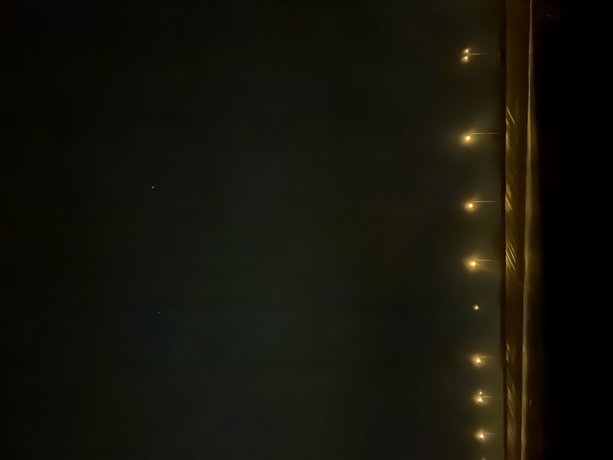 The image shows a dark night scene. Along the right side of the frame, a line of illuminated streetlights is visible, casting a faint golden glow on a vertical surface. The left side of the image is dominated by a dark expanse, possibly the night sky, with one or two tiny points of light that could be stars. The location is unidentified without further evidence.