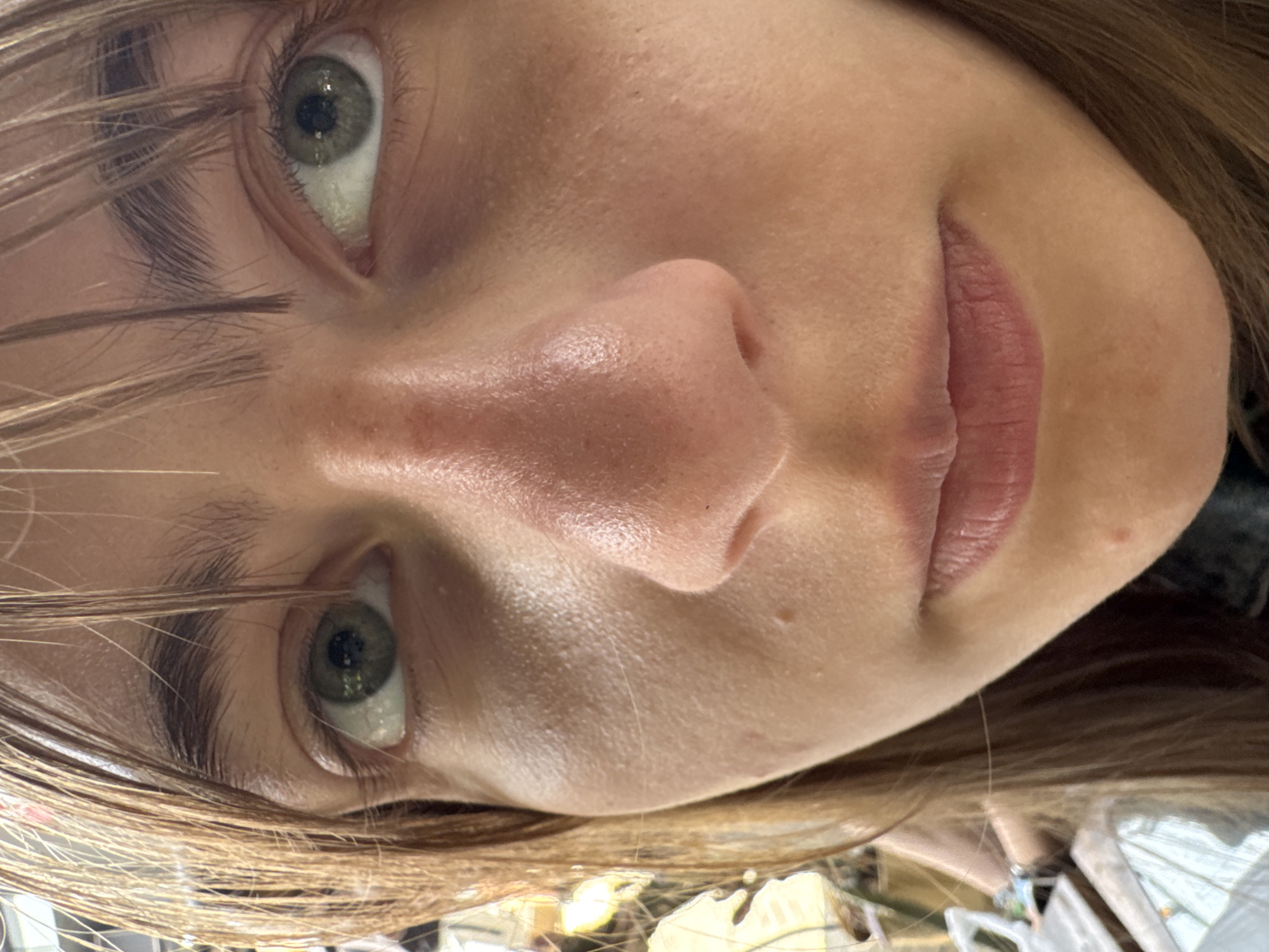 A close-up, slightly angled portrait of a person's face, likely female, with light skin and brown hair partially obscuring the left eye. The person has green eyes, visible pores on the nose and cheeks, and pink lips. The background is out of focus and indistinct. The location and city are Unknown.