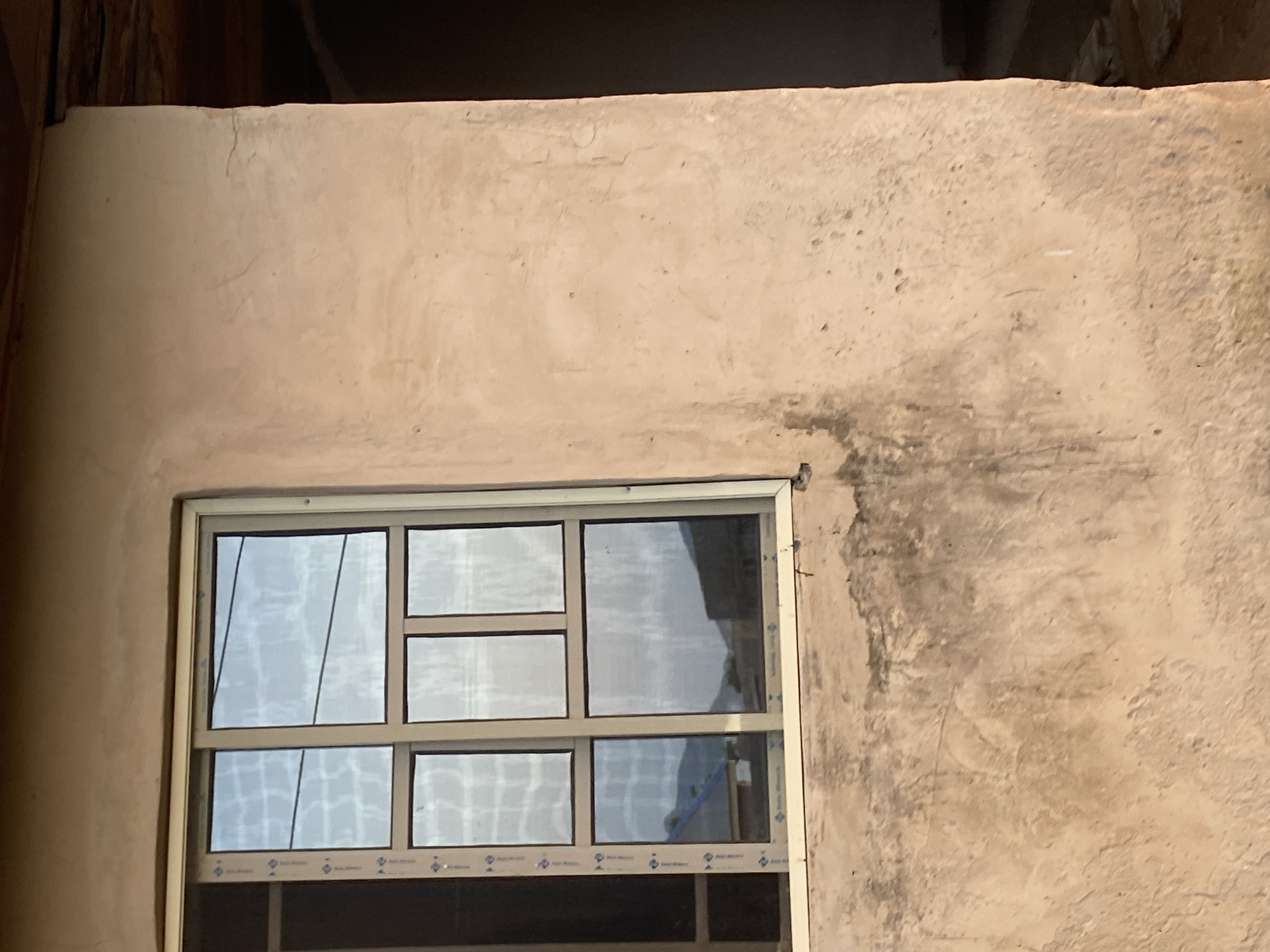 The image shows a close-up of a window on a rough, beige-colored stucco wall. The window has a light-colored frame and is divided into six panes. The glass reflects the sky and some buildings with sloped roofs. The wall itself shows signs of wear, with some darker staining and what appear to be small cracks in the stucco. To the right of the window, the wall transitions to a darker, shadowed area, revealing a glimpse of what might be an interior space with wooden beams visible at the top. The scene appears to be located in Jauro Yinu, Nigeria, and depicts an exterior view of a building under construction or in a state of disrepair.
