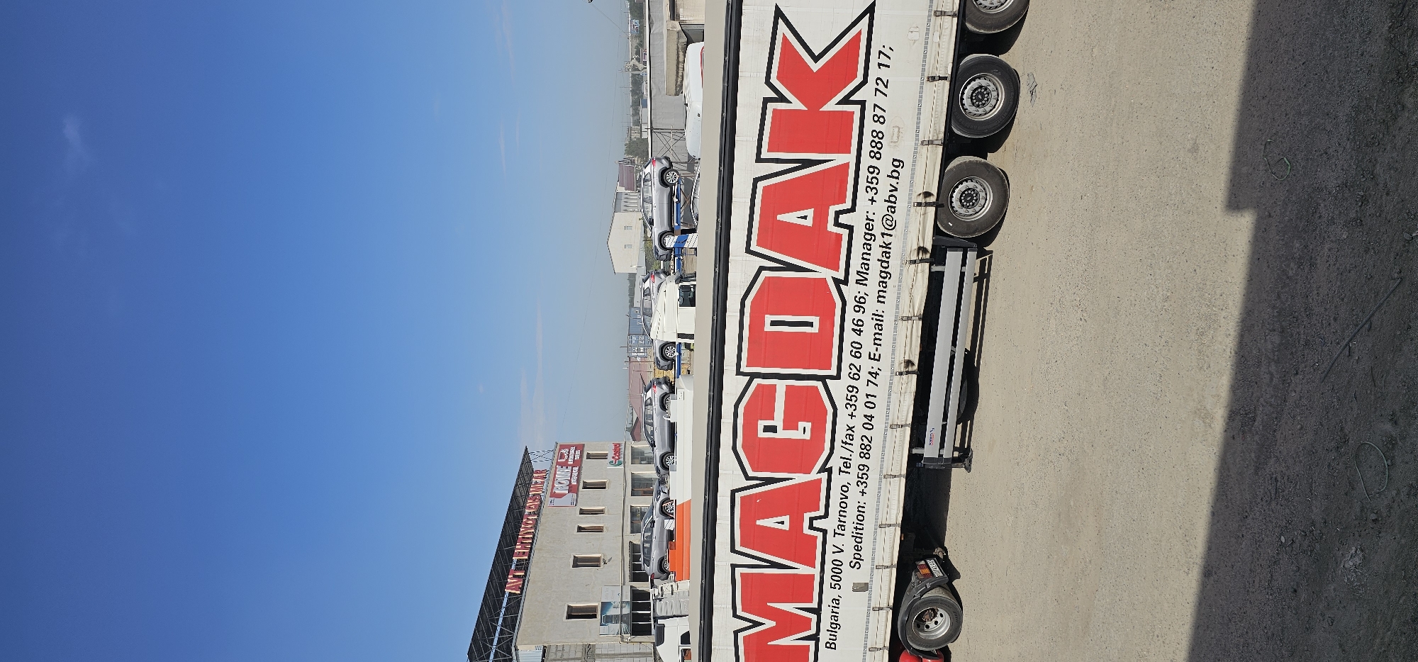 The image shows a white semi-trailer truck with red lettering on its side that reads "MAGDAK". The truck is parked on a dusty, outdoor lot. In the background, there is a building with a sign that says "ANTI-EROKAY PLAY NEAR" and "ROWIYE". Several silver cars are parked in front of the building, some of them on elevated platforms. The sky is clear and blue. The scene appears to be an automotive dealership or repair shop. The location is not specified as Sheyhancha, Uzbekistan, nor is there any indication of this in the image.