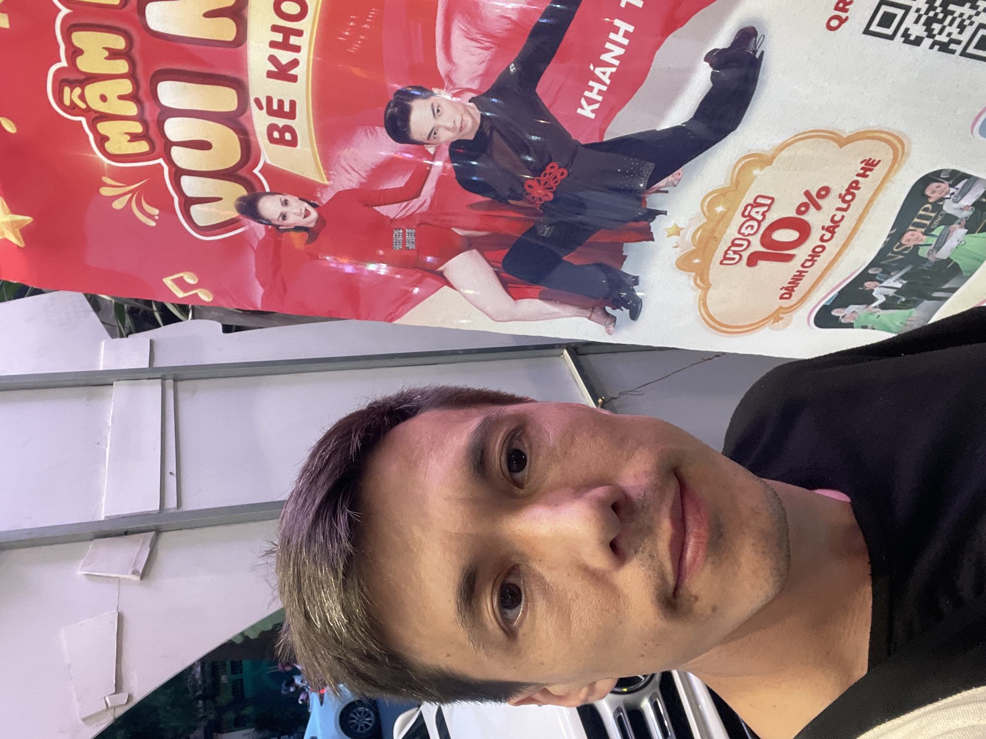 This is a selfie taken by a man in Thủ Đức, Vietnam. He is standing in front of a large red banner advertising a children's program called "Mầm Nụi Bé Khỏe" which translates to "Healthy Buds and Toddlers". The banner features images of two dancers, a woman in a red outfit and a man in a black dance costume. To the right of the banner, there is a promotional element offering a "10% discount for summer classes". The man in the selfie is looking towards the camera with a neutral expression. The lighting suggests it might be daytime, possibly outdoors given the hints of greenery and vehicles in the background. The overall impression is a casual, everyday moment captured in a public or commercial space.
