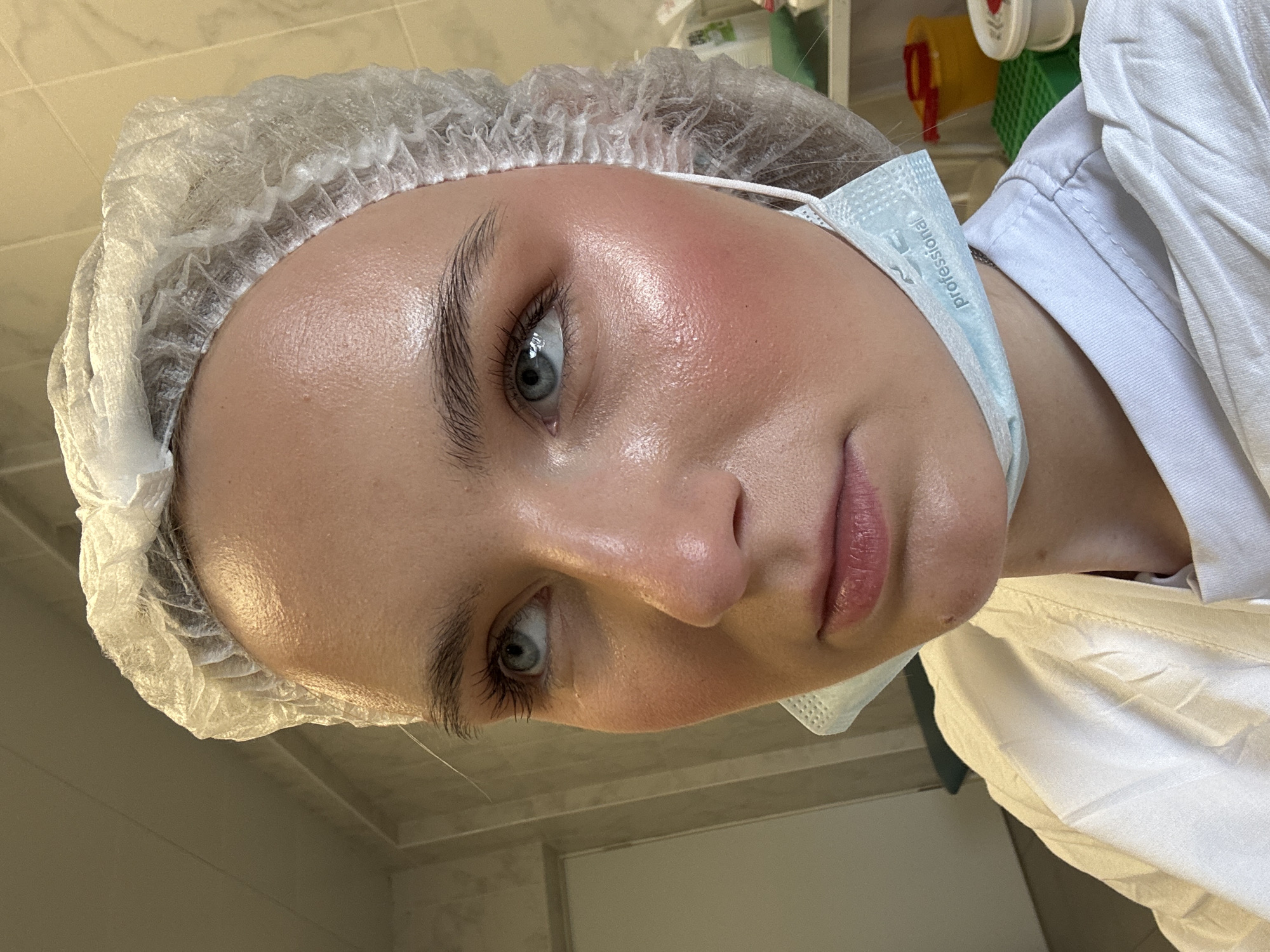 A person with pale skin, blue eyes, and a hairnet on their head is wearing a light blue surgical mask and a white medical gown. The mask is pulled down below the nose. The person is looking at the camera. The background appears to be a medical or clinical setting. There is no evidence of the location being Saint Petersburg, Russia.