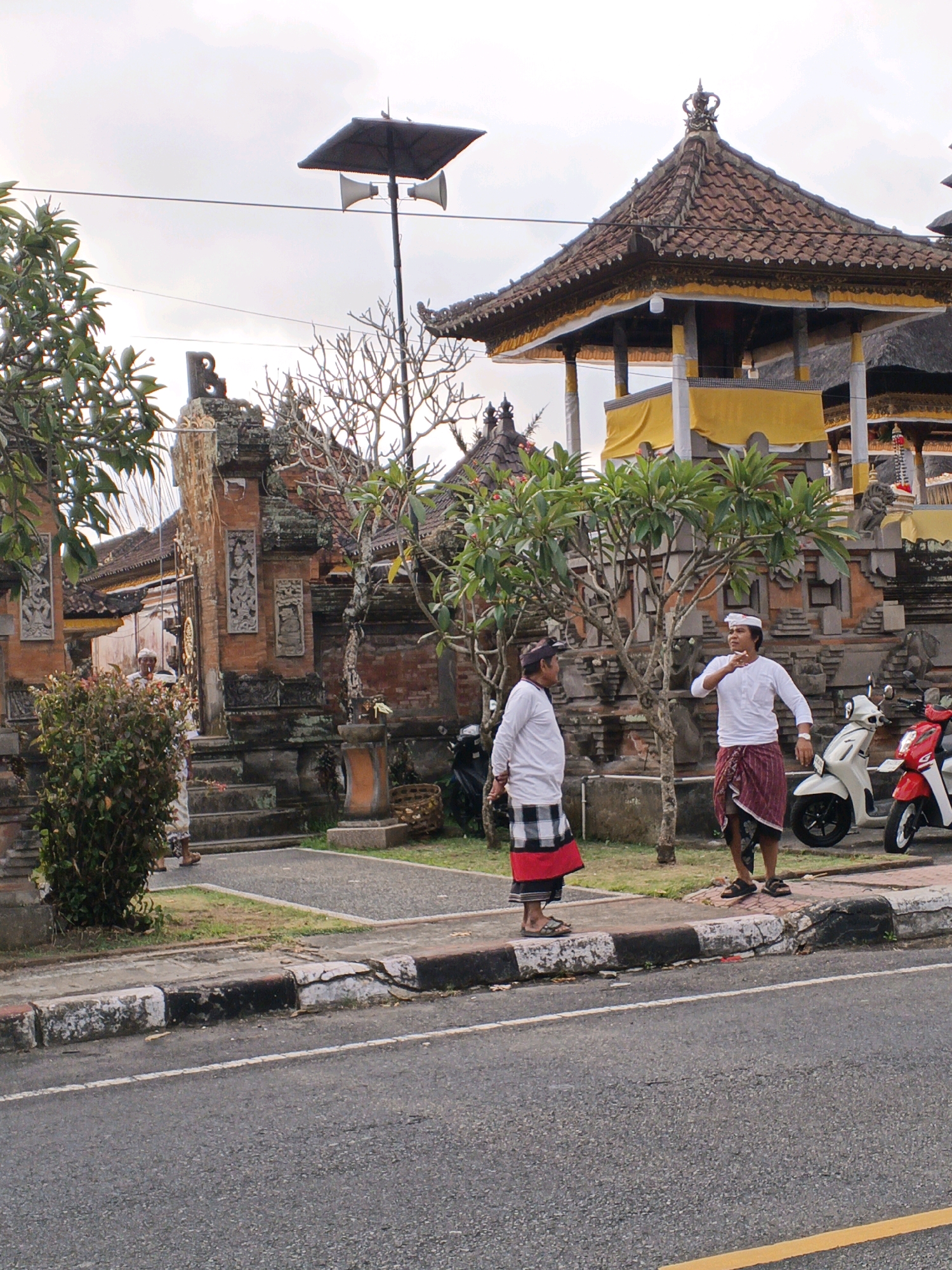 The image captures a street scene in Blahbatuh, Indonesia, featuring traditional Balinese architecture and two men in traditional attire. The foreground shows a paved road with a cracked concrete curb, separating it from a pathway.
On the pathway, two men are positioned near a stone wall and a building. The man on the left, wearing a white shirt and a black and red checkered sarong, stands with his back slightly turned to the camera. The man on the right, also in a white shirt, wears a maroon and black patterned sarong. He appears to be gesturing with his left hand. Behind them, a lush green tree stands prominently.
The background reveals the intricate stonework of a Balinese temple complex, characterized by brick walls, ornate carvings, and tiered roofs covered in clay tiles. A large, covered structure with a crown-like finial, adorned with yellow fabric, is a notable feature. Two motorcycles, one white and one red, are parked on the right side of the pathway. Overhead, a black speaker system is mounted on a pole against a cloudy sky. The overall atmosphere suggests a cultural setting within the town of Blahbatuh.