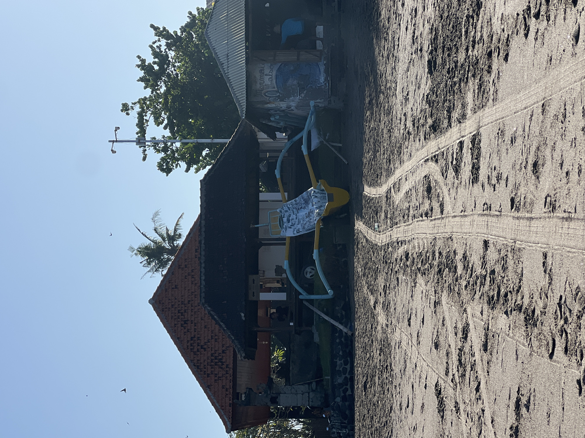 The image depicts a beach scene in North Kuta, Indonesia, characterized by dark sand and clear blue skies. In the foreground, tire tracks are visible on the sand, suggesting vehicle activity. Several small birds are in flight in the upper portion of the frame. Dominating the mid-ground are two structures: a larger building with a reddish-tiled roof and an open-sided covered area, and a smaller, more rudimentary structure with a corrugated metal roof. A traditional outrigger canoe, painted yellow and white with blue outriggers, is positioned between these buildings. The canoe is resting on a wooden stand. The larger building appears to have an entrance with visible steps, and possibly a person inside. The smaller building has graffiti on its exterior and an open doorway where a person in a blue shirt is visible. Trees and vegetation are present behind the structures, and a lamppost stands tall to the right. The overall impression is a somewhat rustic beachside location.