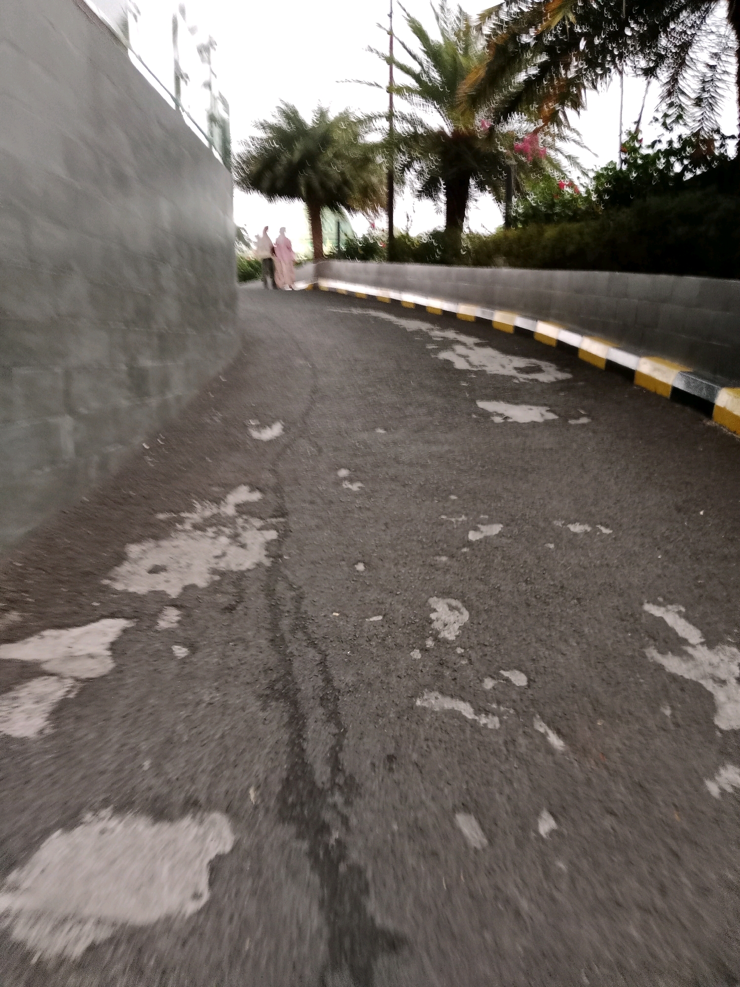 The image depicts an outdoor scene, likely a pathway or road with a textured asphalt surface. The surface is uneven, with lighter-colored patches that could indicate wetness or staining. A grey, textured wall runs along the left side of the frame, appearing to curve away from the viewer. To the right of the pathway, a low curb with alternating yellow, black, and white painted sections defines the edge. Beyond the curb, there is dense green foliage and palm trees. Two individuals, appearing as indistinct figures in light-colored clothing, are walking away from the camera on the pathway. The sky is overcast and bright. The scene has a slightly blurred or motion-affected quality.