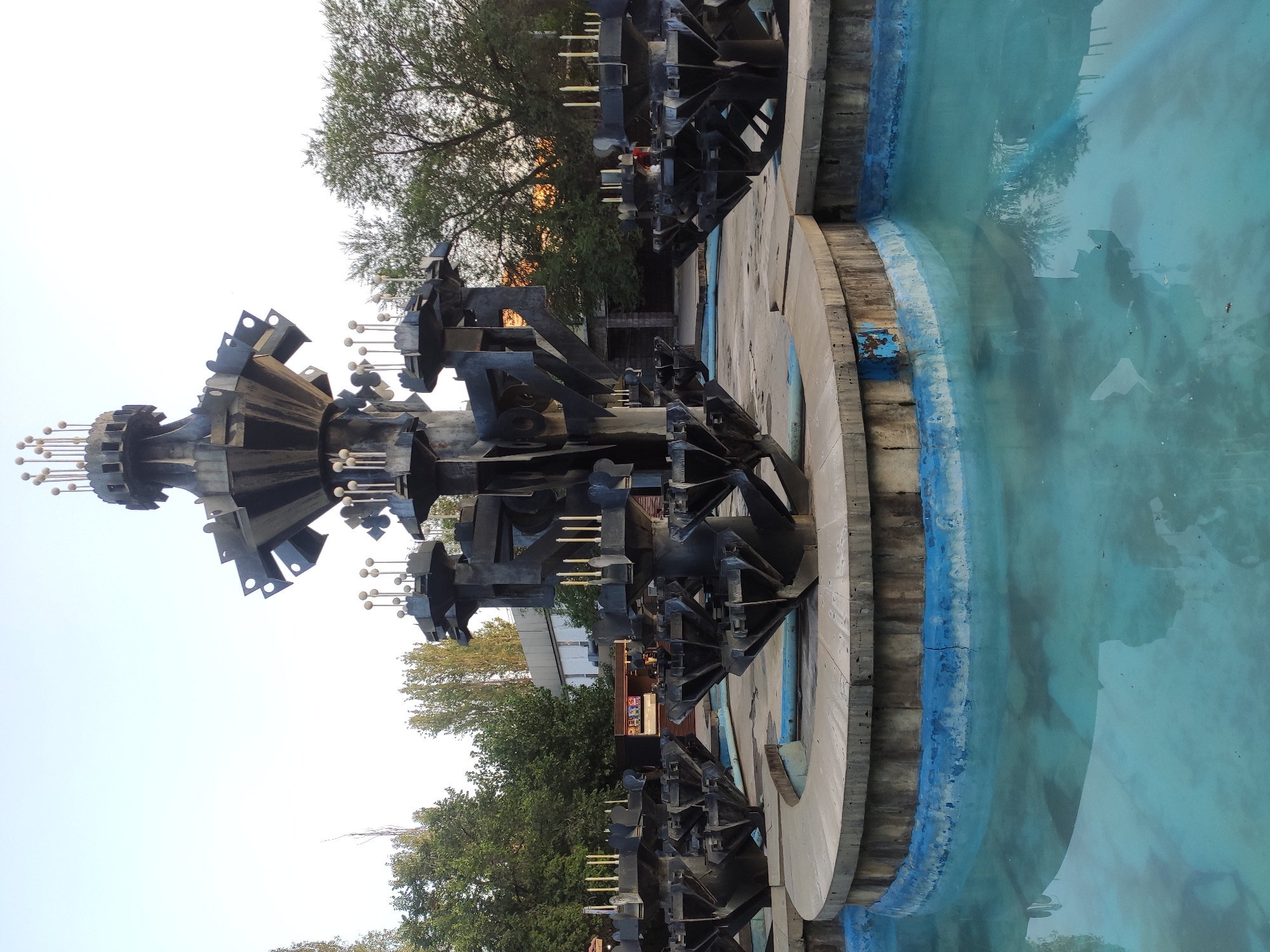 This is a photograph of the "Singing Fountain" located in Republic Square in Yerevan, Armenia. The fountain, a large, multi-tiered, dark metal sculpture with spherical elements, is the central subject. It stands in a circular pool filled with turquoise water. The base of the fountain is made of concrete with a blue-painted rim, which shows signs of wear and chipping. Several smaller, similar-looking sculptural elements surround the main structure, also within the water. In the background, there are trees with green foliage and a clear sky with a hint of sunset colors. A building with windows is visible behind the fountain and trees. The overall scene suggests an outdoor public space, likely a park or plaza, in the late afternoon or early evening. There are no people visible in the immediate foreground or interacting with the fountain.