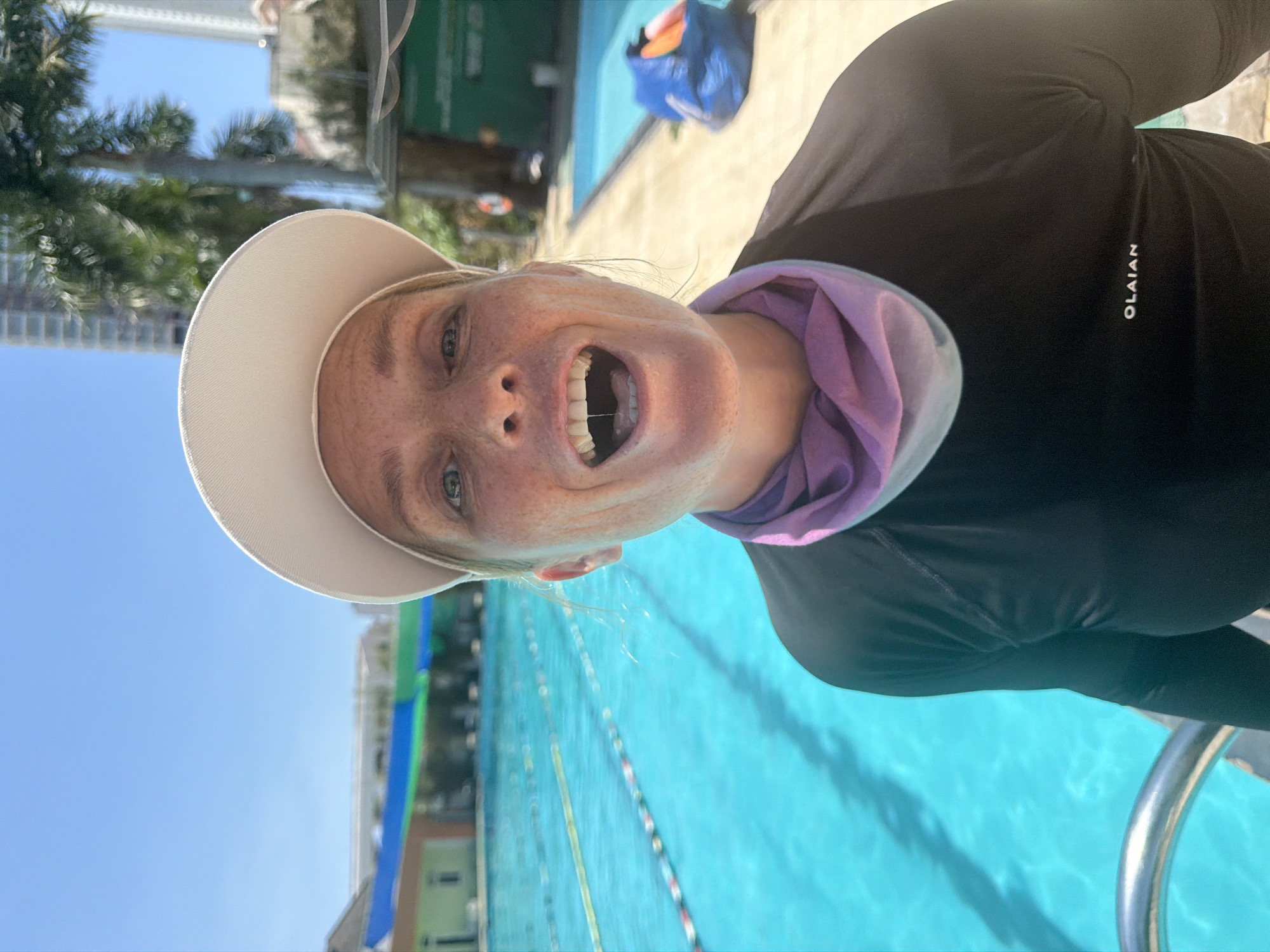 The image shows a close-up, self-portrait of a woman with freckles, wearing a white hat and a black long-sleeved shirt with a purple scarf around her neck. She is smiling with her mouth open, revealing her teeth. Her eyes are blue and she has blonde hair. The setting appears to be outdoors, by a swimming pool with clear blue water and lane markers. In the background, there are trees, buildings, and a bright blue sky, suggesting a sunny day. The woman is looking directly at the camera, conveying a cheerful and energetic emotion. The text "OLAIAN" is visible on her shirt. The scene is likely a resort or public swimming facility. The time of day appears to be daytime, given the bright sunlight and clear sky. The location context provided is Nha Trang Ward, Vietnam.