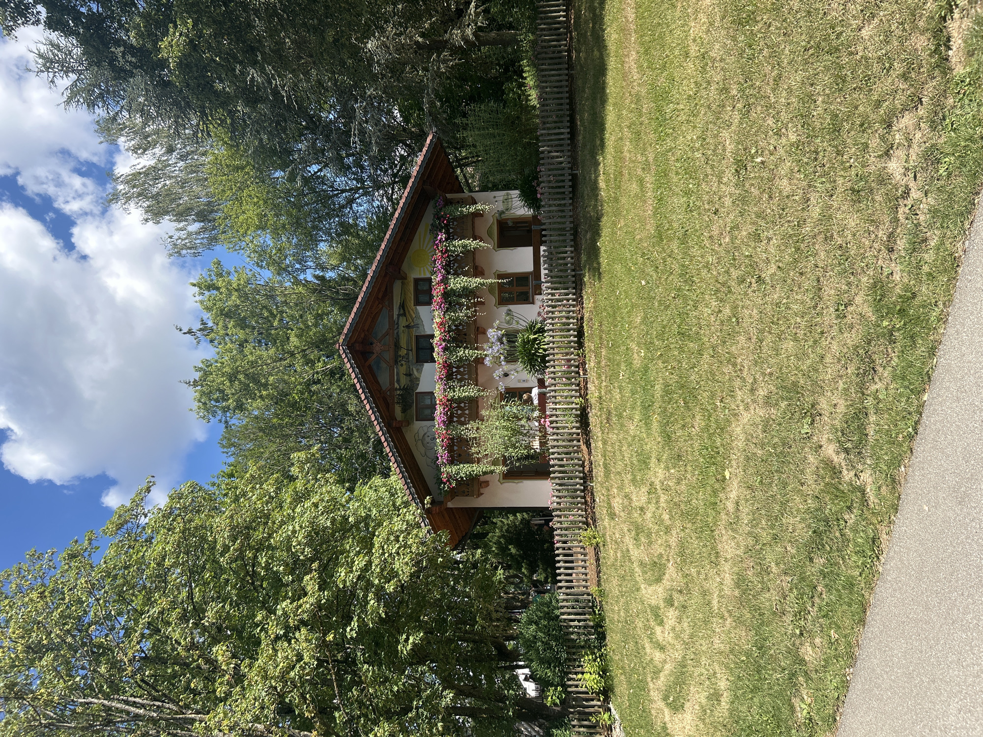 The image depicts a charming Bavarian-style house located in Reisbach, Germany. The house is painted a light color with decorative murals on its facade, featuring a sun and figures. A prominent feature is the balcony adorned with an abundance of pink and red flowers. A wooden picket fence encloses the property, separating it from a grassy yard. The yard shows signs of dryness in some areas, suggesting a sunny climate. Tall green trees surround the house, providing a lush backdrop. The sky above is a bright blue with scattered white clouds. A paved path is visible in the lower left corner of the image. The scene appears to be a serene, picturesque residential setting in a rural or semi-rural area of Reisbach.