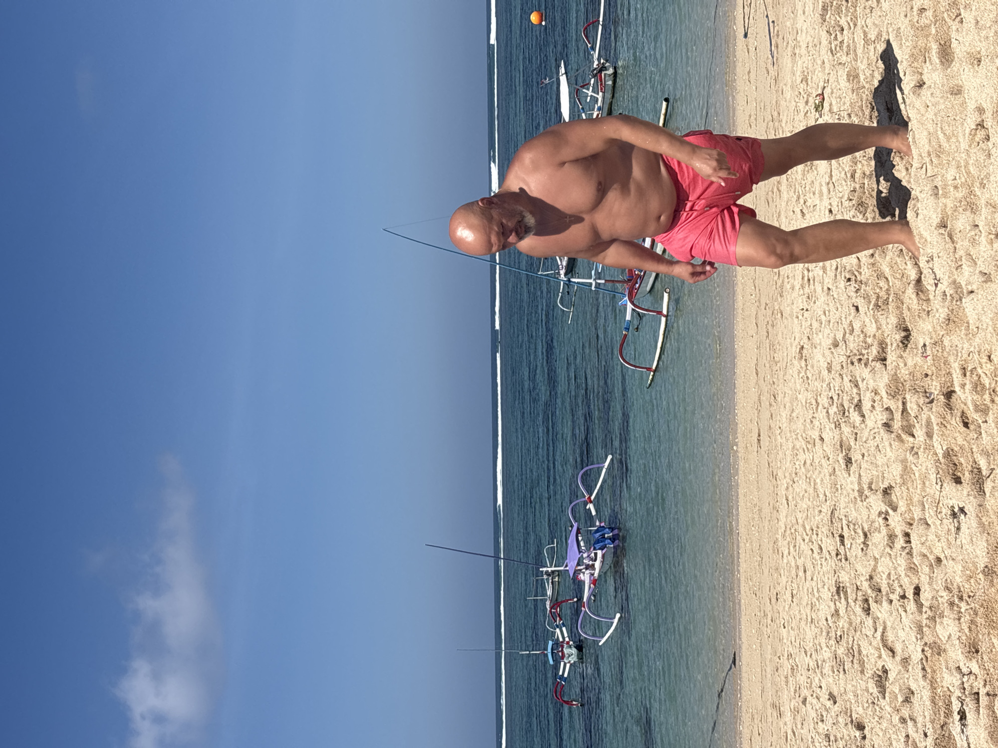 A bald, heavyset Caucasian man wearing red swim trunks walks on a sandy beach. He has a light beard and is looking down. In the background, the turquoise ocean meets the horizon under a clear blue sky with a few wispy clouds. Several traditional outrigger boats are moored in the shallow water. The scene is likely Kuta Selatan, Indonesia, known for its beaches and these types of boats.