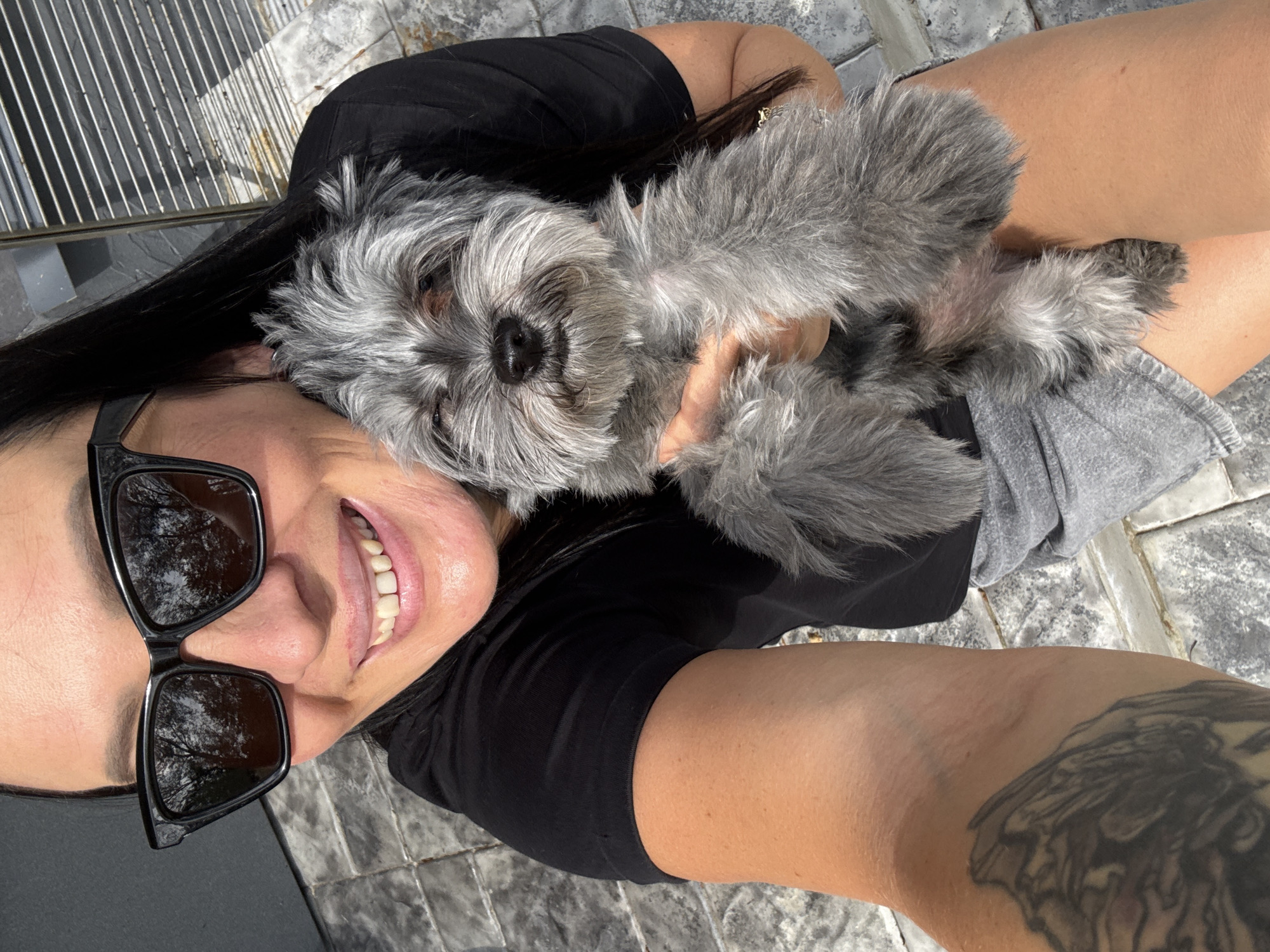 In a close-up, outdoor selfie, a woman with long dark hair and sunglasses smiles broadly, holding a small, gray, fluffy dog. The woman is wearing a black t-shirt and gray shorts. The dog appears to be a Schnauzer mix. The background consists of a paved patio with gray tiles and a slatted metal gate. The image was taken in Chaikovsky, Russia, on a sunny day.
