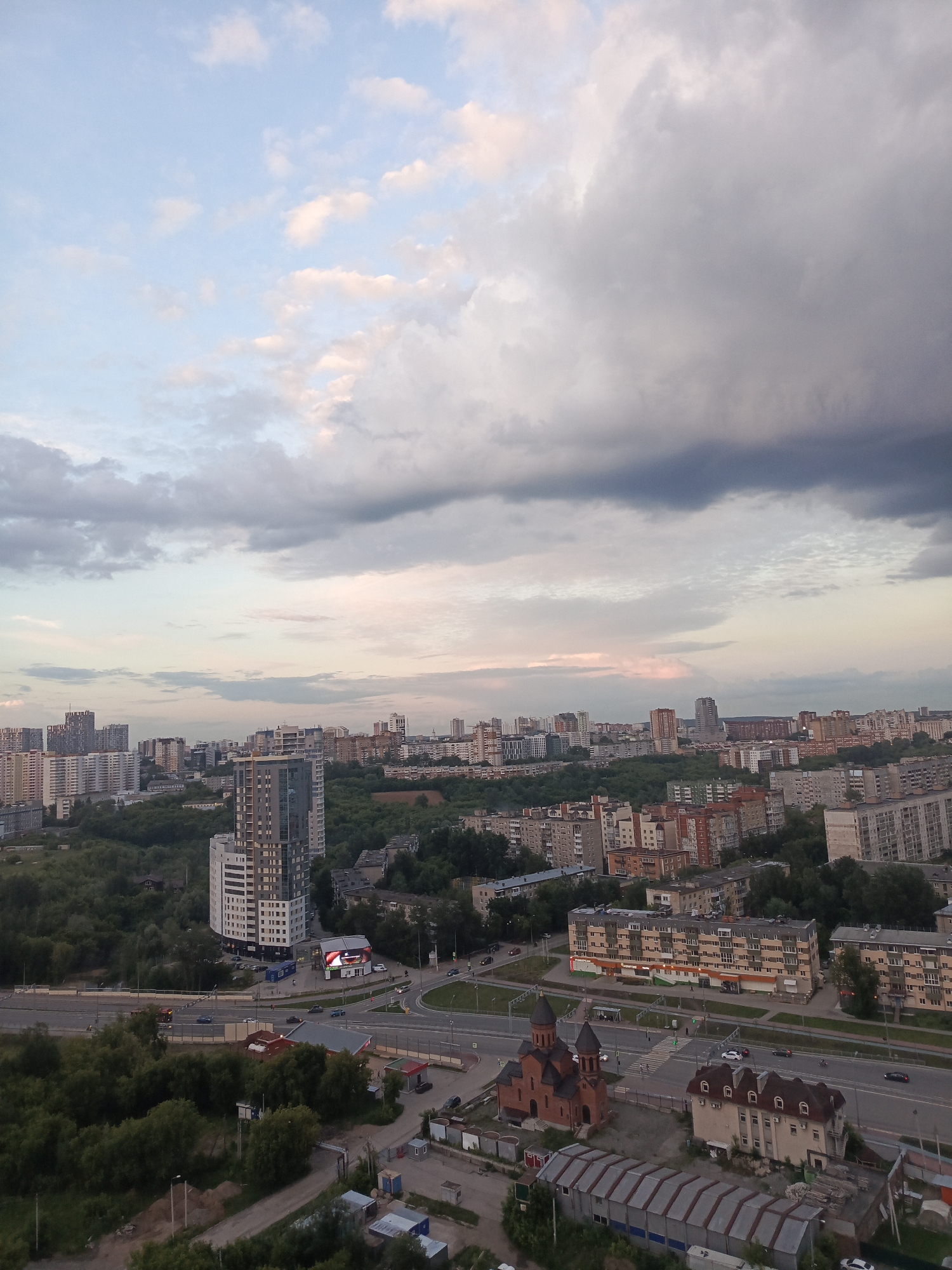 The image shows a panoramic view of a city with many apartment buildings and some green spaces. In the foreground, there are trees and some industrial-looking structures. A red brick church with dark domes is visible, along with a tan building with a dormer roof. A road runs through the scene with several cars driving on it. In the middle ground, there are more apartment buildings and a densely wooded area. A tall, modern apartment building stands out. In the background, the city continues with more residential buildings extending to the horizon. The sky is partly cloudy with a mix of gray and pinkish hues, suggesting either sunrise or sunset. This is a scene from the city of Perm, Russia.