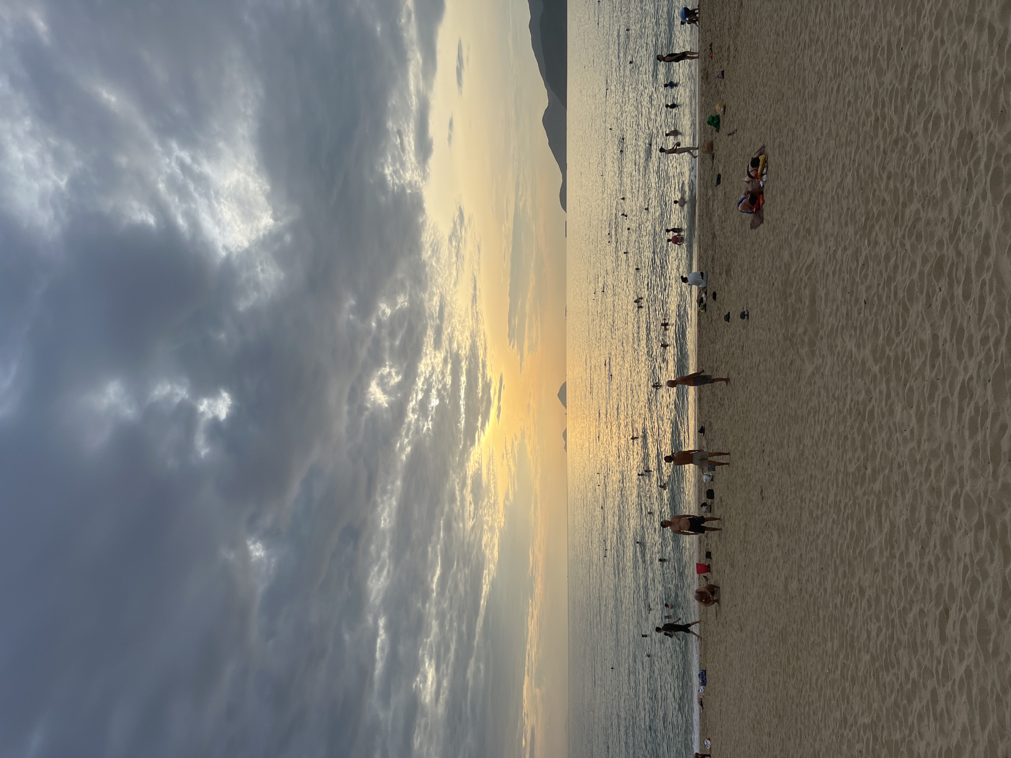 This image depicts a wide, sandy beach in Nha Trang, Vietnam, during what appears to be late afternoon or early evening. The sky is filled with dramatic, dark clouds that are breaking to reveal the warm glow of the setting sun, casting a golden hue over the horizon and the calm ocean.
Numerous people are scattered across the beach and in the water. On the sand in the foreground, several individuals are standing or sitting, some in swim trunks, suggesting they are enjoying a day at the beach. Further back, closer to the water's edge, more people are visible, with some wading or swimming in the sea. A significant number of individuals are present in the ocean, indicating a popular spot for recreation.
The water is relatively calm, with gentle waves lapping at the shore. In the distance, faint outlines of islands or peninsulas can be seen against the brightly lit horizon. The beach itself is composed of fine, light-colored sand, with no visible structures or significant debris in the immediate foreground. The overall atmosphere is one of a relaxed, end-of-day beach scene.
