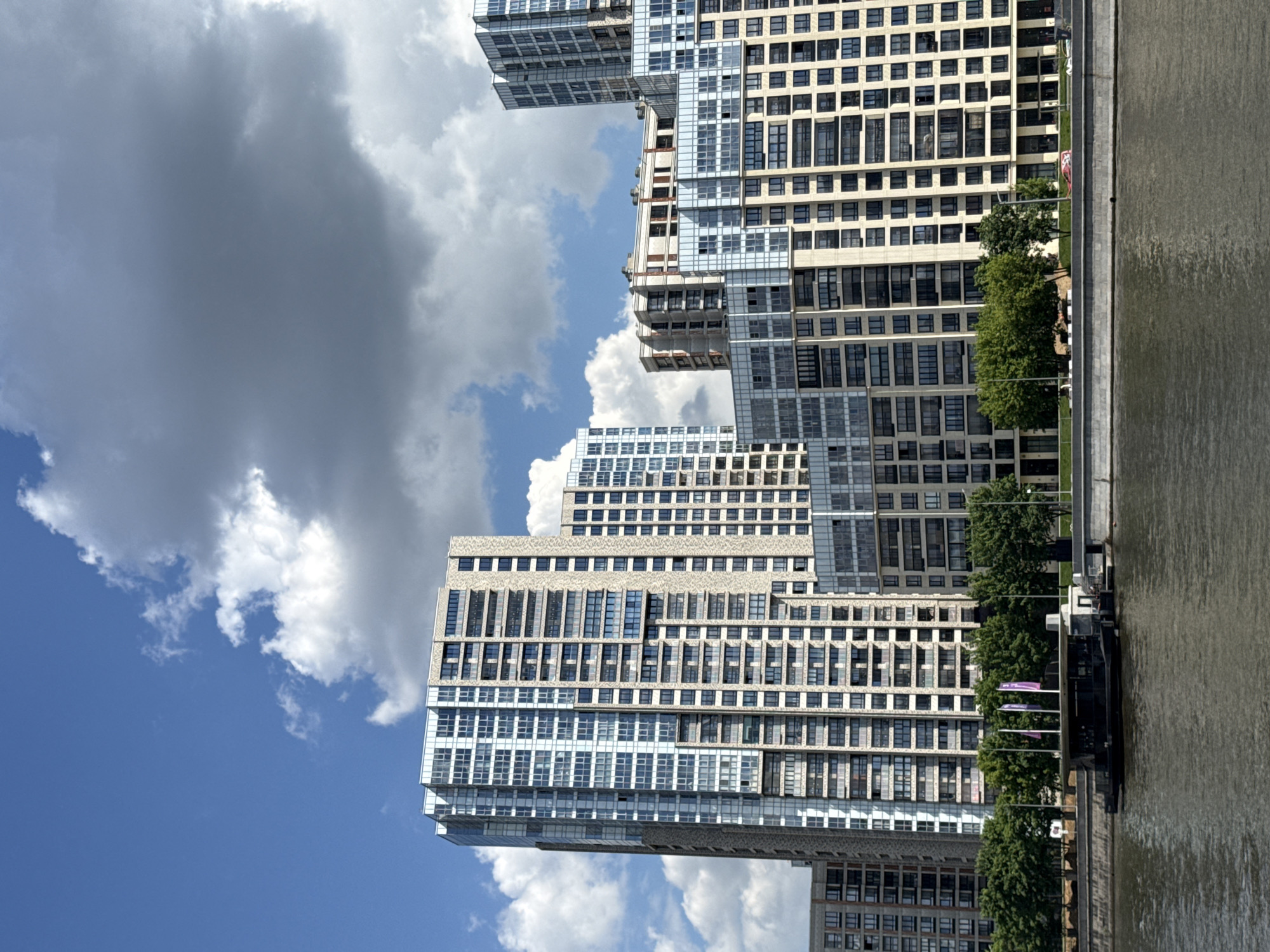 This media file captures a vibrant modern waterfront scene in Moscow, Russia. The image is oriented with the sky at the top, buildings on the left, and a river on the right.
The scene features several imposing, contemporary multi-story buildings lining the riverbank. These structures appear to be residential, characterized by numerous square and rectangular windows set into light-colored facades, interspersed with sections of glass curtain walls. The architecture is modern and geometric, with some buildings exhibiting tiered designs and cantilevered elements, creating an dynamic skyline.
Separating the buildings from the river is a well-maintained embankment, featuring a line of lush green trees that provide a natural border. A wide river occupies the right side of the frame, its surface showing reflections of the sky and subtle ripples, indicating movement. Moored along the embankment, a dark-colored boat or barge is visible towards the lower right. Further along the embankment, three purple flags on poles add a splash of color.
The weather is bright and clear, with a vivid blue sky dotted with fluffy white clouds and some larger, darker grey clouds, suggesting a sunny daytime. There are no people visible, lending a serene atmosphere to the urban landscape. The overall impression is one of modern development along a scenic waterway. No discernible text is visible.