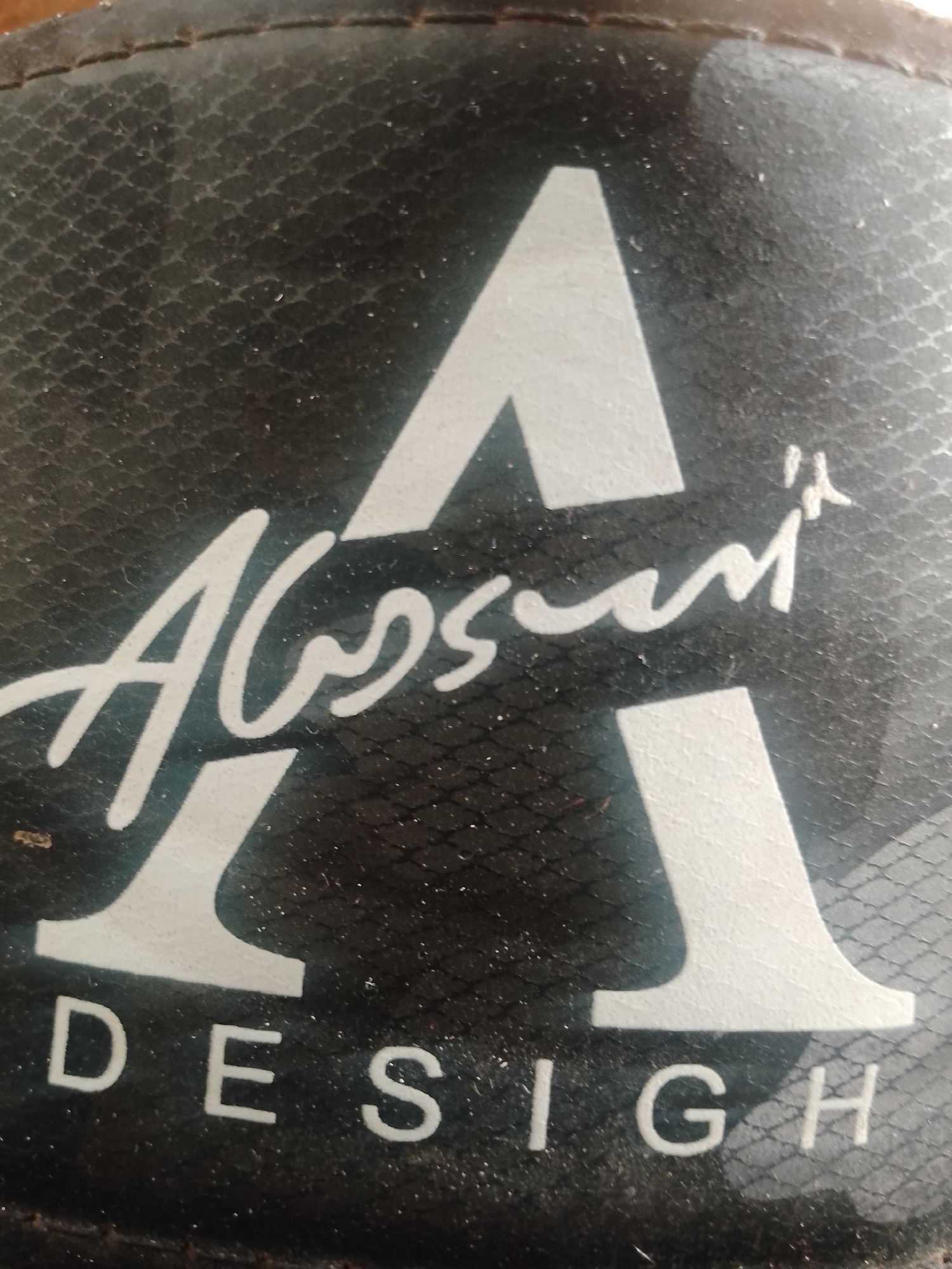The image shows a close-up of a black textured surface with a large white stylized letter "A" superimposed. Below this, in a cursive white font, is the word "Absun". Further down, in block white letters, is the word "DESIGN". The surface appears to be made of a synthetic material with a subtle diamond pattern. The lighting casts shadows and highlights on the textured surface and the lettering. There is no indication of the image's location within Jalingo, Nigeria, or any other specific environmental details.