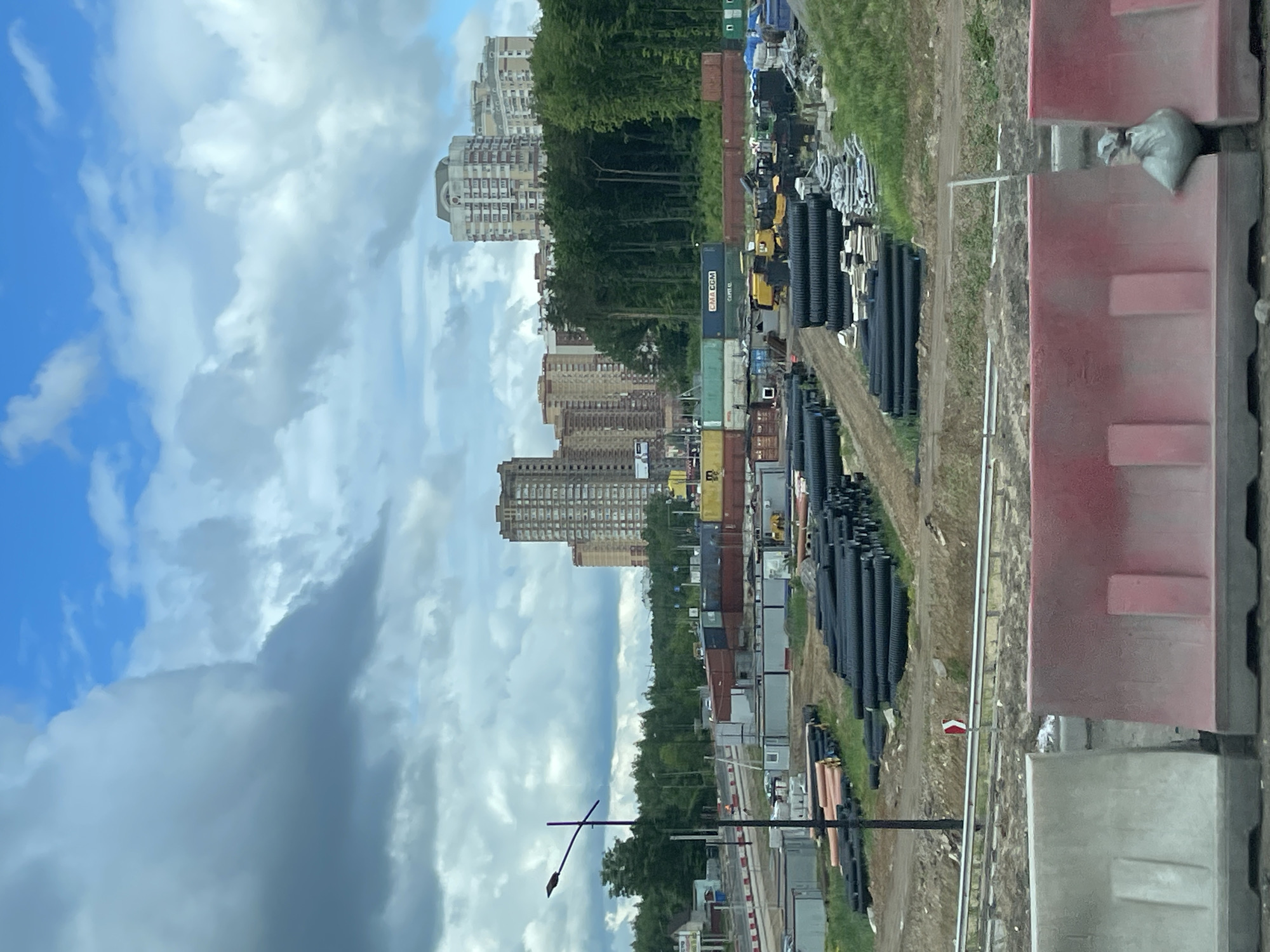 The image shows a view in Novodrozhzhino, Russia. The scene includes residential high-rise buildings, trees, stacks of black plastic pipes, a row of shipping containers and temporary office units, and construction barriers. The sky is cloudy with patches of blue. A lamppost is visible. The overall impression is of an urban environment with construction activity or material storage near residential areas.