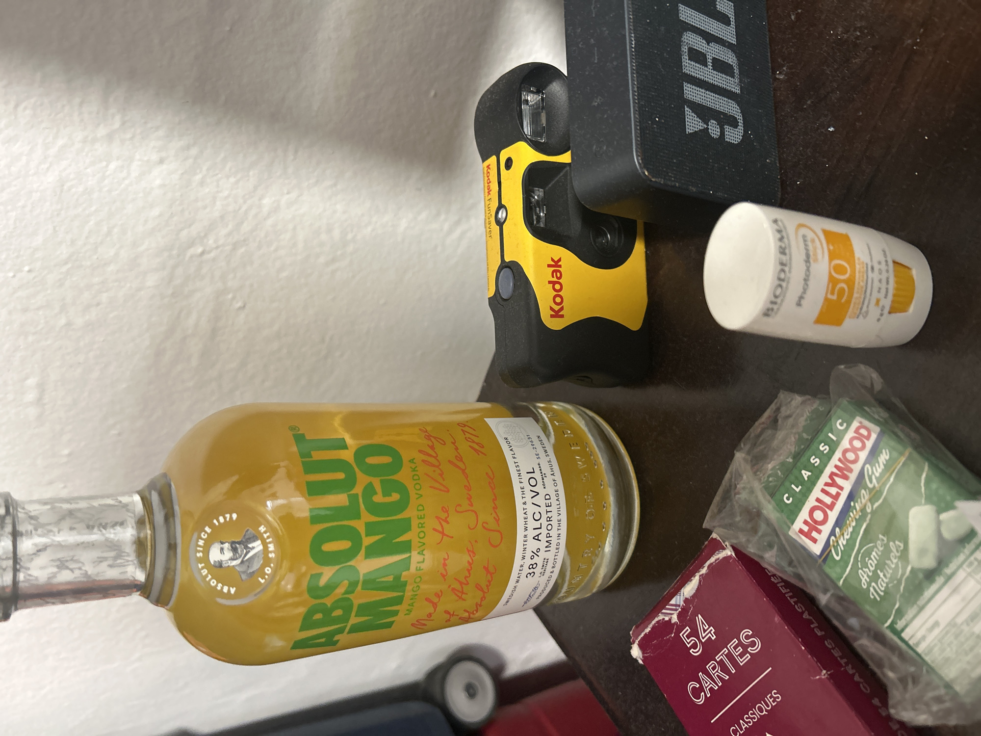 The image shows a collection of items on a dark wooden surface. The main object is a bottle of Absolut Mango flavored vodka, lying horizontally. Next to it is a yellow and black Kodak disposable camera and a black JBL speaker. A tube of Bioderma sunscreen with SPF 50 is visible near the speaker. There is also a partially visible pack of Hollywood chewing gum in a green box, alongside a red box labeled "54 Cartes Classiques". There are two small, toy-like wheels visible beneath the vodka bottle. The background includes a plain white wall.
There is no mention of Sainte-Anne, France, in the image content.