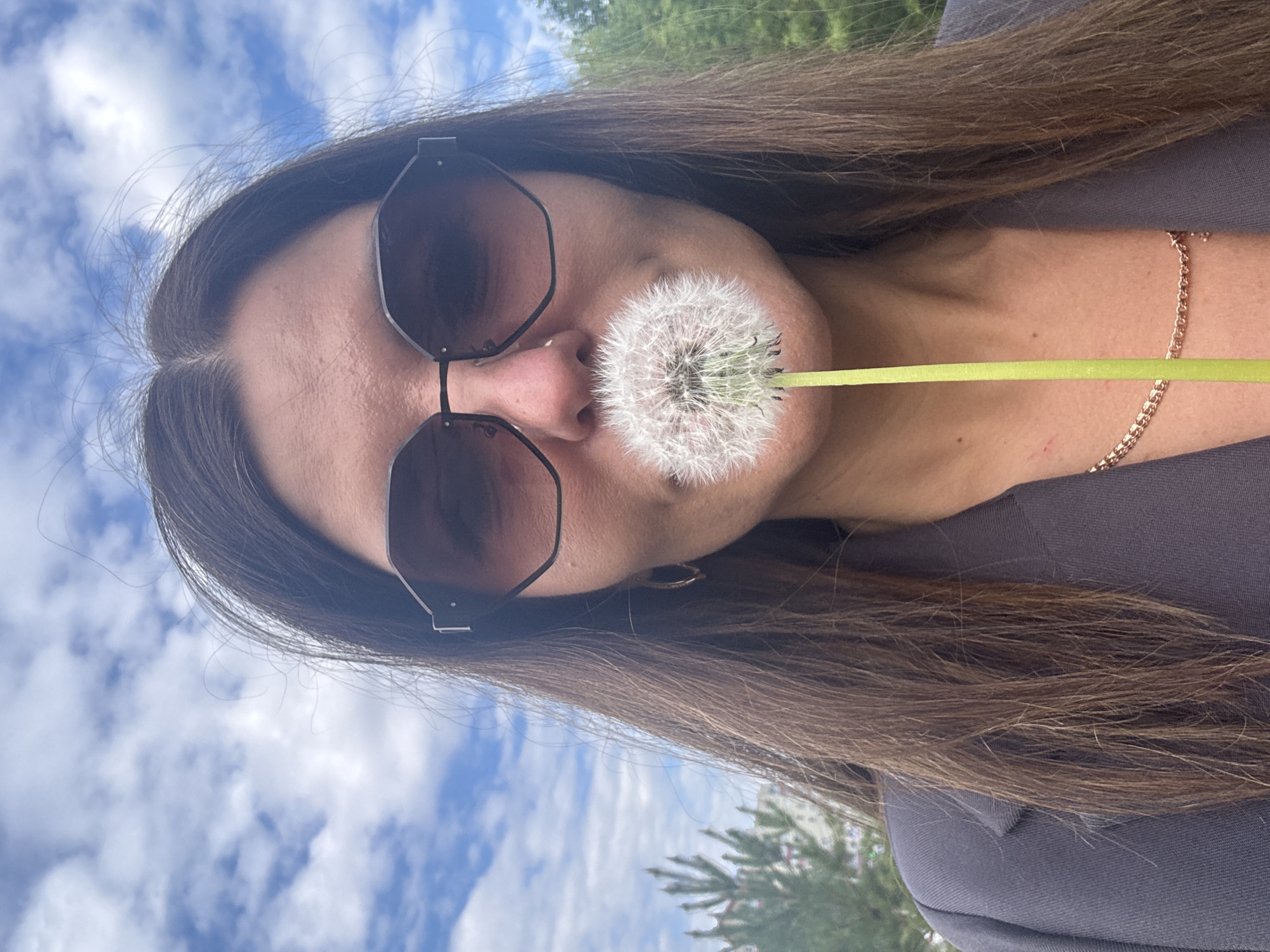 The image displays a close-up, horizontally oriented shot of a person outdoors. The individual's head is on the left, with long, dark brown hair extending across the frame to the right. They are wearing dark, hexagonal-shaped sunglasses that obscure their eyes. A fully bloomed dandelion seed head is held just below their nose, resting against the upper lip area, with its bright green stem pointing to the right.
The person is dressed in a dark grey top or jacket. A thin, golden chain necklace is visible around their neck, and a matching golden chain bracelet is worn on their right wrist. The background is divided; the upper portion reveals a bright blue sky with scattered white clouds, while the lower portion shows blurred green foliage, indicative of trees or bushes. The natural lighting suggests a clear and sunny daytime. The activity involves a playful interaction with the dandelion. There is no visible text in the image.