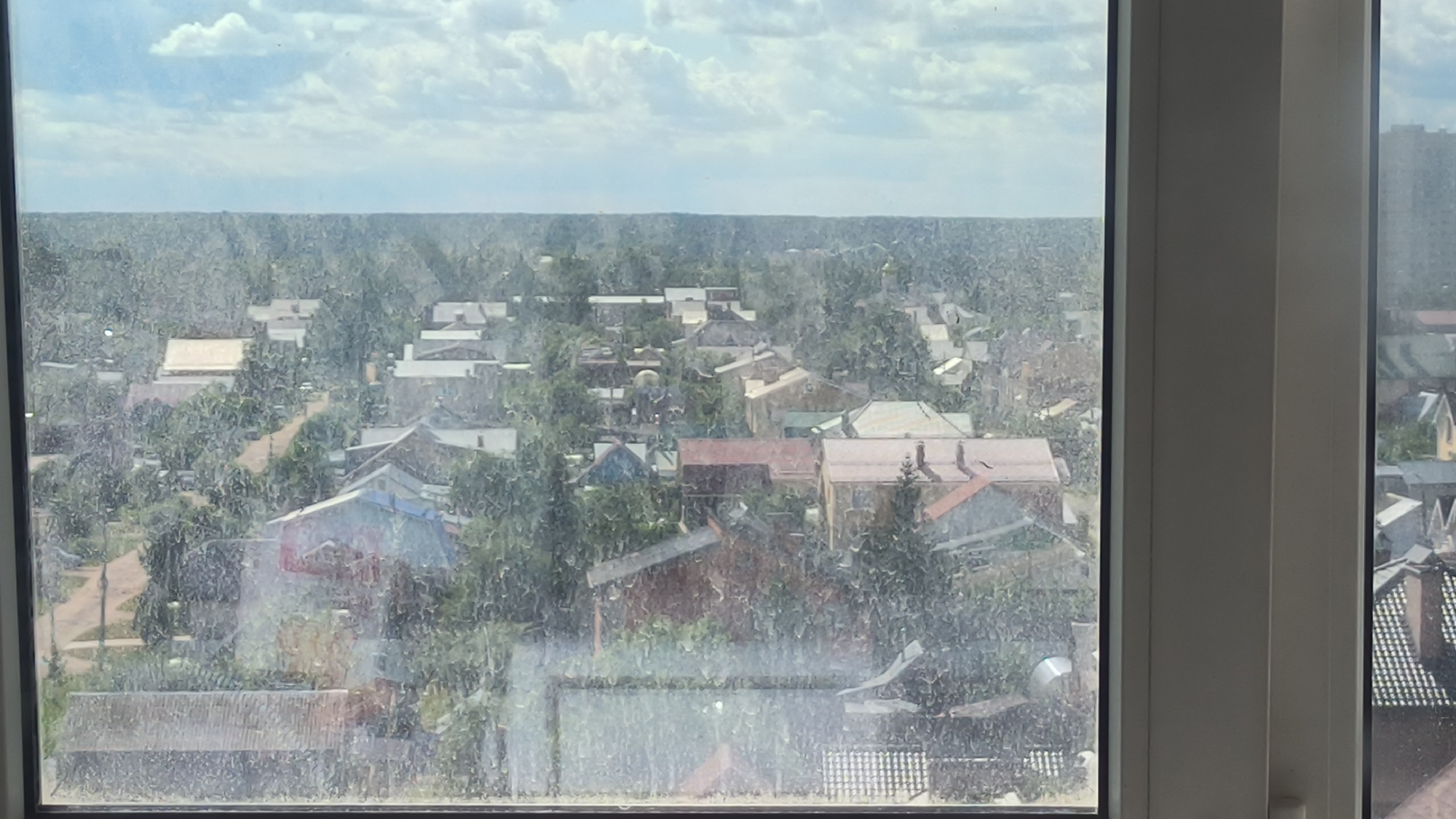 The image shows a view of a residential area in Kovrov, Russia, seen through a dirty window. The scene depicts a cluster of houses with red, brown, and white roofs, interspersed with numerous trees. In the distance, a faint horizon is visible. A tall, modern building can be seen on the far right side of the frame. The overall impression is that of a sunlit, overcast day.