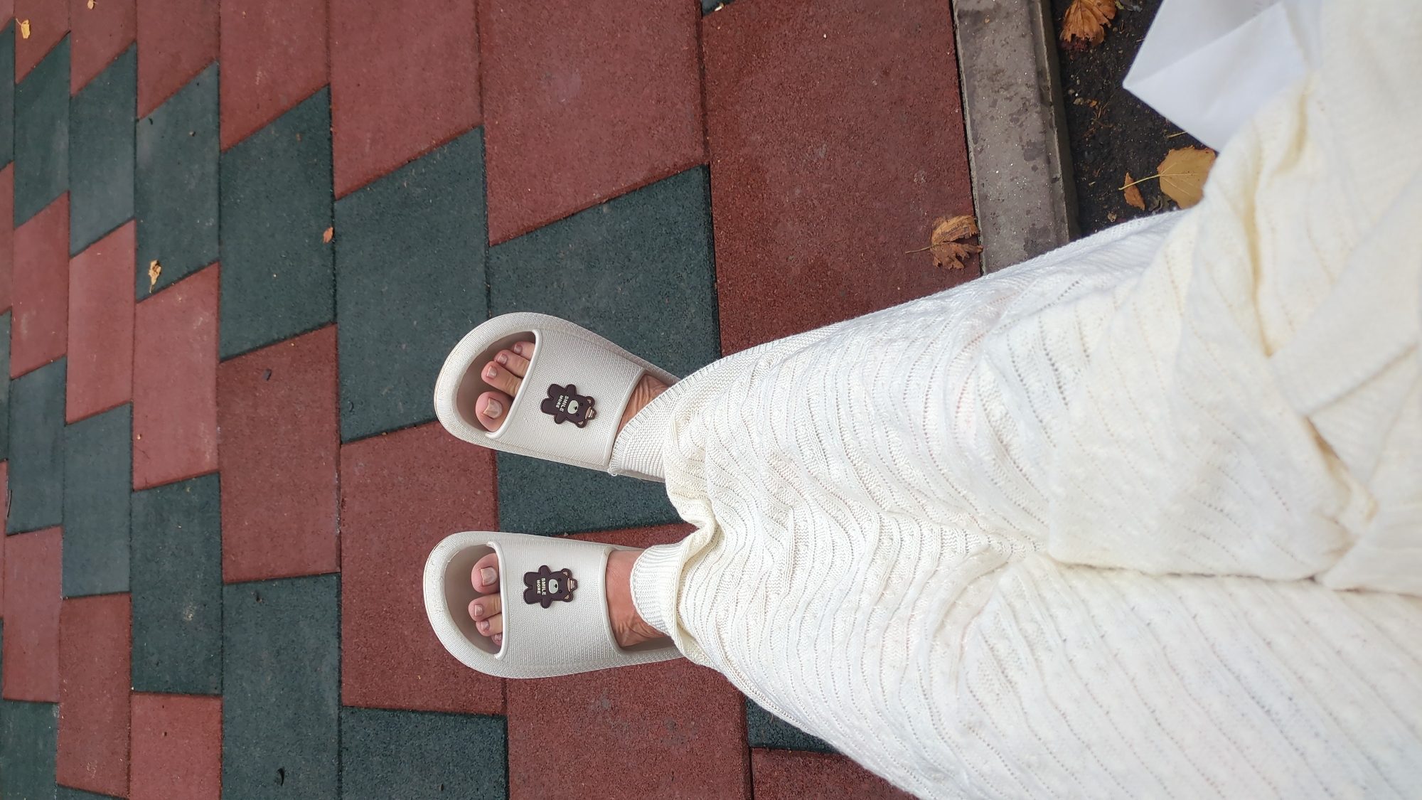 A pair of feet, wearing white slide sandals with a bear design, are visible against a backdrop of red and dark gray paving stones. The feet are partially covered by a cream-colored knitted garment. Fallen leaves are scattered on the ground. The scene appears to be outdoors in an urban setting, potentially a park or public walkway in Chișinău, Moldova, given the paving style.