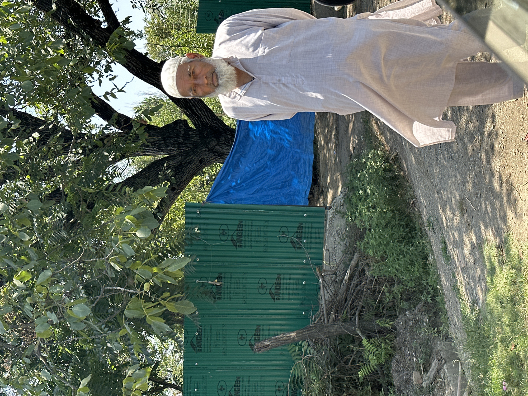 In the city of Vessu, India, an elderly man with a white beard and a white skullcap stands in the foreground. He is wearing a light-colored, loose-fitting tunic and pants. Behind him, a large tree with green leaves provides shade. A bright blue tarp is visible behind the man and to his left, and a green corrugated metal fence is in the background. The ground is dirt and gravel, with some green vegetation growing on the left side of the image. The lighting suggests it is daytime.