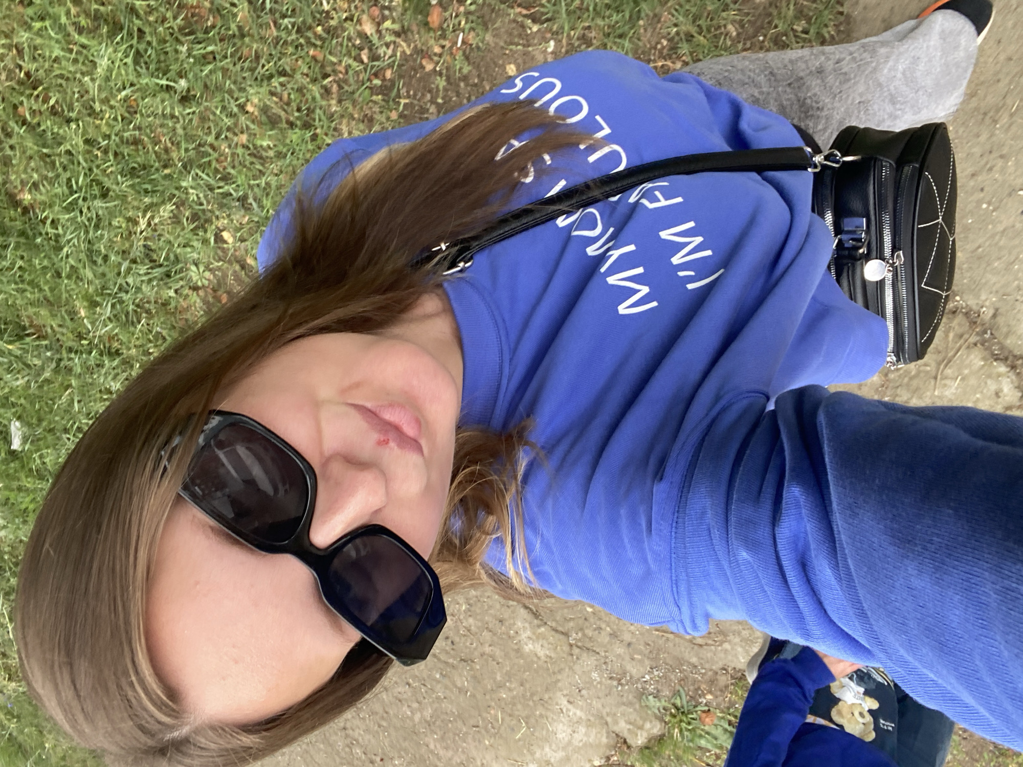 A selfie taken from a high angle shows a woman standing outdoors in what appears to be a residential or park area. She has long brown hair and is wearing large black sunglasses, a bright blue sweatshirt with white text that reads "MY MOM SAID I'M FABULOUS," and grey sweatpants. A small black crossbody bag with silver hardware is slung over her shoulder. Her lips are slightly parted, and there appears to be a small blemish or mark on her upper lip. The background consists of green grass and a paved path. The lighting suggests it is daytime, likely overcast. There is no visible indication of the city of Noviy, Russia, or any specific landmarks.