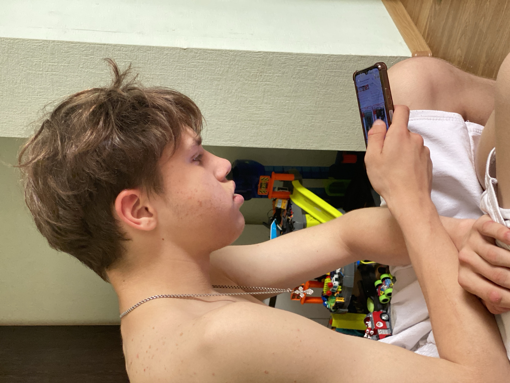 A shirtless, light-skinned male with short brown hair is indoors and looking at a smartphone he is holding in his right hand. He is wearing a silver necklace with a cross pendant. He is partially covered with a white cloth. In the background is a toy race track with several colorful cars. The location cannot be determined.