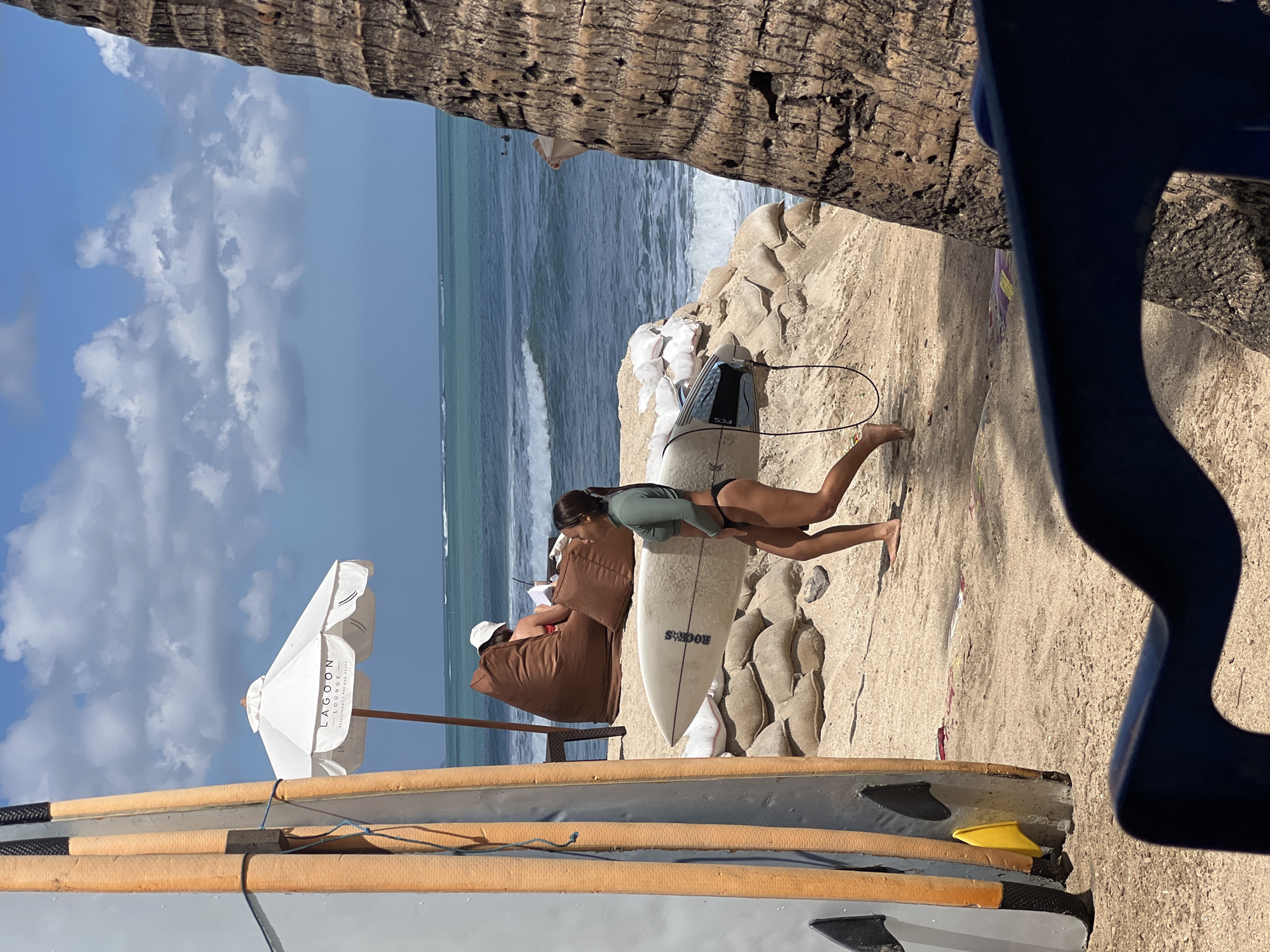 In Kuta, Indonesia, on a sandy beach, a woman in a green long-sleeved surf top and black bikini bottoms carries a surfboard and walks towards the ocean. She is on the right side of the frame, with a palm tree trunk partially obscuring her. In the background, a person sits on a large brown beanbag chair under a white umbrella that reads "Lagoon Lounge," seemingly reading a book. Several surfboards are propped up on the left side of the image, and sandbags are stacked on the beach. The ocean is visible in the background with gentle waves, and the sky is blue with scattered clouds.