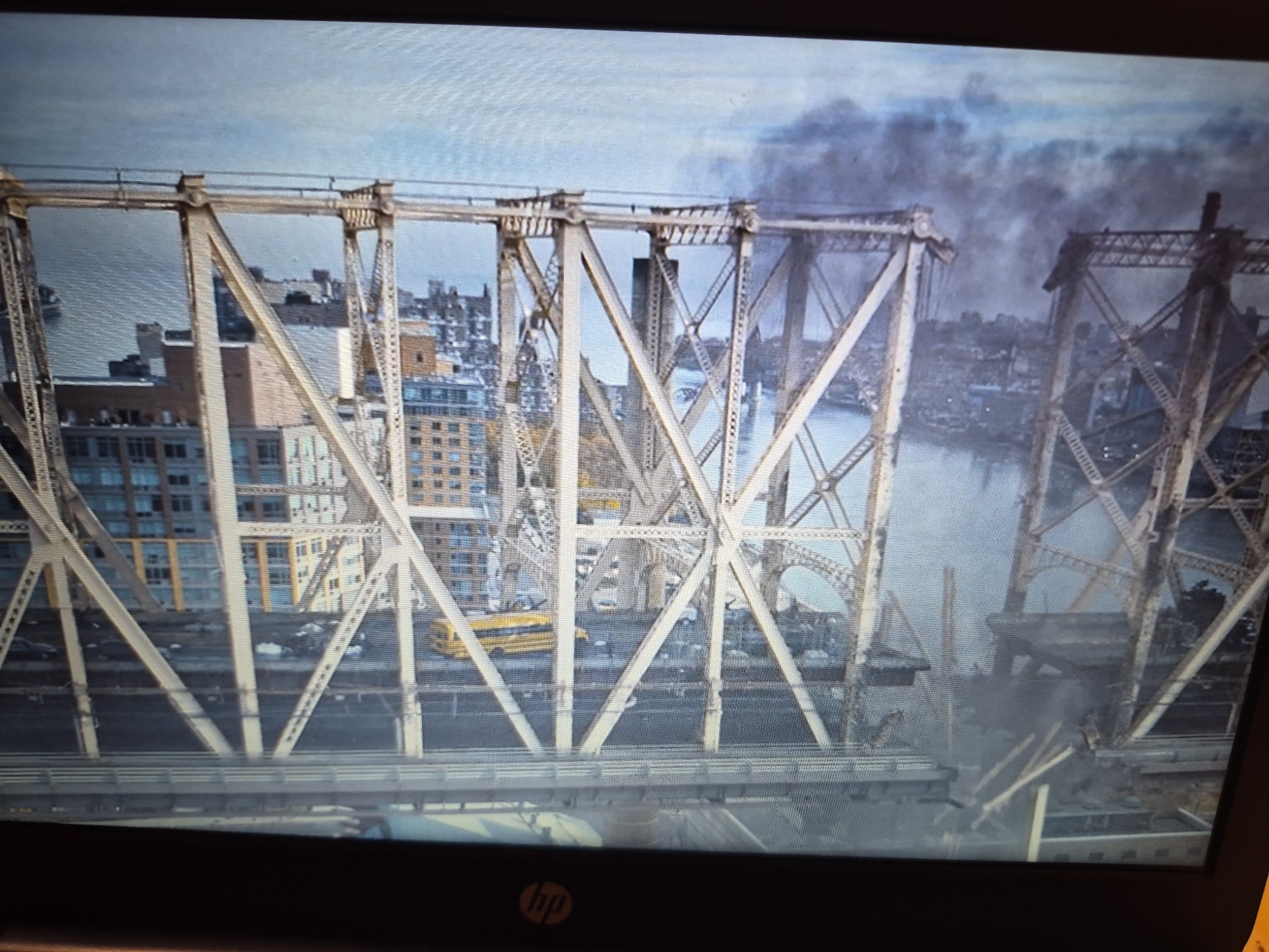 The image depicts a partially destroyed bridge structure in a city, with smoke rising in the background, suggesting a recent catastrophic event. The bridge appears to be made of steel trusses and is spanning a body of water, likely a river. Buildings are visible on either side of the river, indicating an urban environment. The bridge itself shows significant damage, with large sections missing or collapsed. Traffic, including cars and a yellow school bus, is visible on the intact portion of the bridge. The sky is overcast with some clouds. The scene is rendered in a slightly grainy, low-resolution aesthetic. There is no discernible location in Sandwell, United Kingdom.
