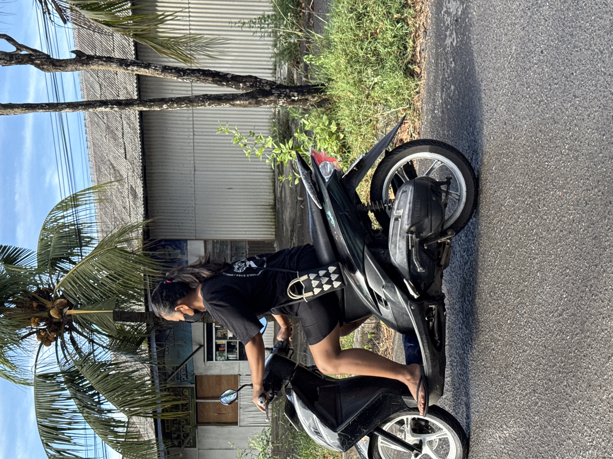 A medium outdoor shot depicts an individual on a black scooter on an asphalt road in Kuta Selatan, Indonesia. The person, with dark hair pulled back, is wearing a black t-shirt, black shorts, and flip-flops, and a black face mask. The scooter is parked on the side of the road. In the background, there's a corrugated metal building with a palm tree to the left. Power lines are visible above. Vegetation lines the side of the road. The setting suggests a tropical or semi-tropical urban environment.