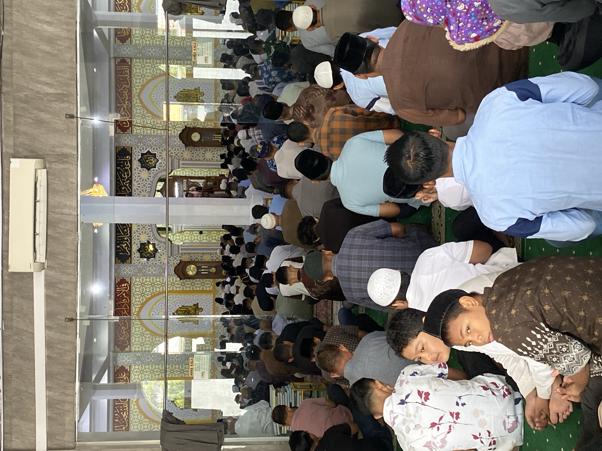 The image depicts the interior of a mosque in Lhokseumawe, Indonesia, likely during a prayer service. A congregation of men and boys are seated on a green carpeted floor, facing the front of the mosque where decorative Islamic calligraphy adorns the walls. Some individuals are looking down, presumably in prayer or contemplation, while others are looking forward.
In the foreground, several young boys are visible. One boy in a white shirt with a floral pattern is seated, with his feet bare. Next to him, a boy wearing a black peci (cap) and a patterned brown shirt is looking towards the camera. Another boy in a white peci is visible to his right.
The mosque interior features ornate architecture with arched doorways and patterned walls in yellow and white. A large grandfather clock stands in the background, along with a sign that reads "AWAS KACA" (Warning: Glass). An air conditioning unit is mounted on the ceiling. The scene conveys a sense of community and religious observance.