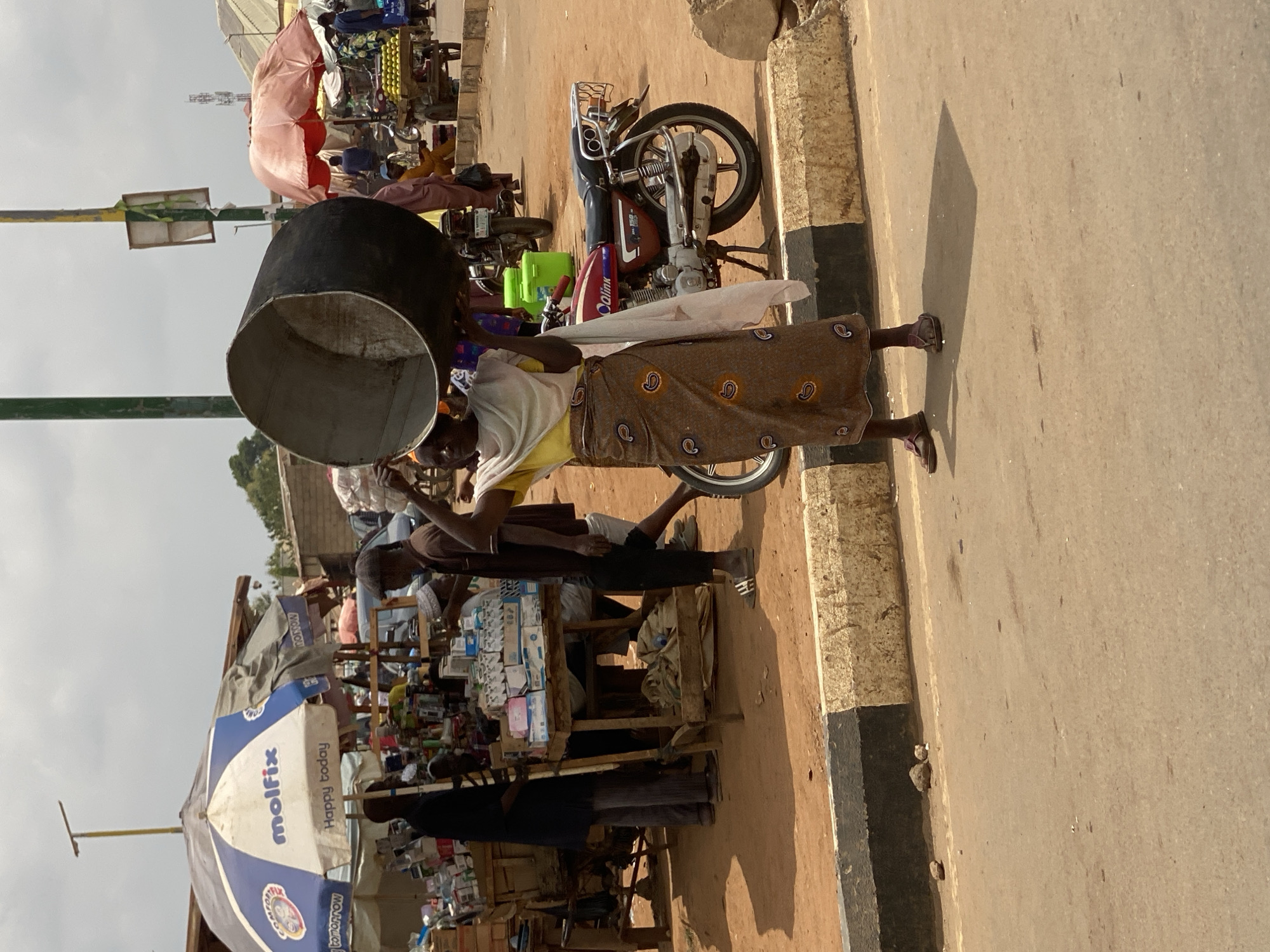 The image captures a vibrant street scene in Kafanchan B, Nigeria, during what appears to be a sunny day, evidenced by strong shadows. In the foreground, a person, likely a woman, is centered, carrying a large, dark, cylindrical container or basin on her head. She is dressed in a light-colored top and a brown patterned skirt or wrap, and wears sandals.
To her right, a red and black motorcycle, with "Qalink" visible on its tank, is parked by the roadside. The road itself is made of asphalt, with a black and white painted curb marking its edge. Behind the primary subject, the street extends into a bustling market area with several stalls. One prominent stall is covered by a blue and white umbrella, which has the brand names "molfix" and "Happy today" clearly printed on it, with "tomorrow" partially visible.
Various goods, appearing as packaged items and boxes, are displayed on the market stalls. Other individuals are present in the background, some engaged in market activities near the stalls. The ground is dry and dusty in areas, consistent with an outdoor market setting. A utility pole with a small rectangular sign is also visible on the left side of the frame. The overall scene depicts daily life in a lively market environment.
