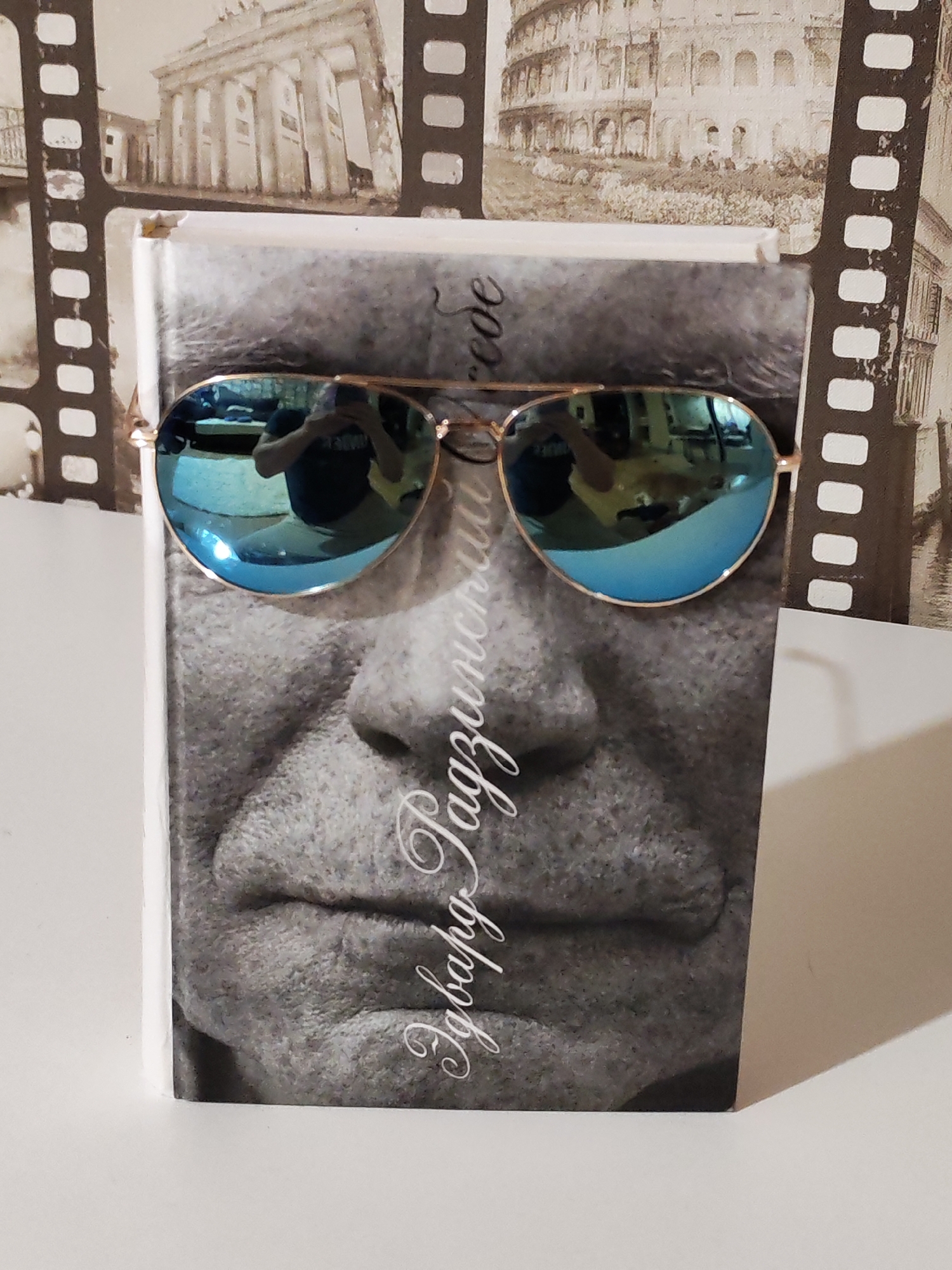 The image shows a book with a black and white portrait of a person. Blue aviator sunglasses are placed on the book, obscuring the eyes of the person depicted. The book is sitting on a white surface. The background is a wall with a film reel themed wallpaper featuring images of European landmarks. The location is likely indoors. The city of Chaikovsky, Russia is not explicitly visible in the image.