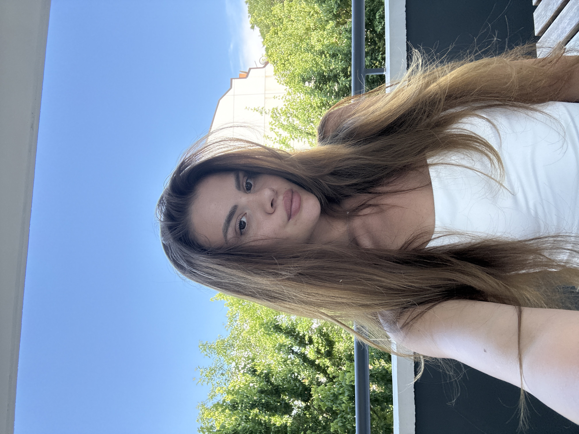 A young woman with long, brown hair is taking a selfie outdoors. She is wearing a white tank top and is positioned on a balcony with lush green trees and a building in the background. The sky is clear blue. The balcony railing is visible in the foreground. The scene appears to be a residential area, likely in the city of Plovdiv, Bulgaria, given the architectural style of the building and the type of vegetation.