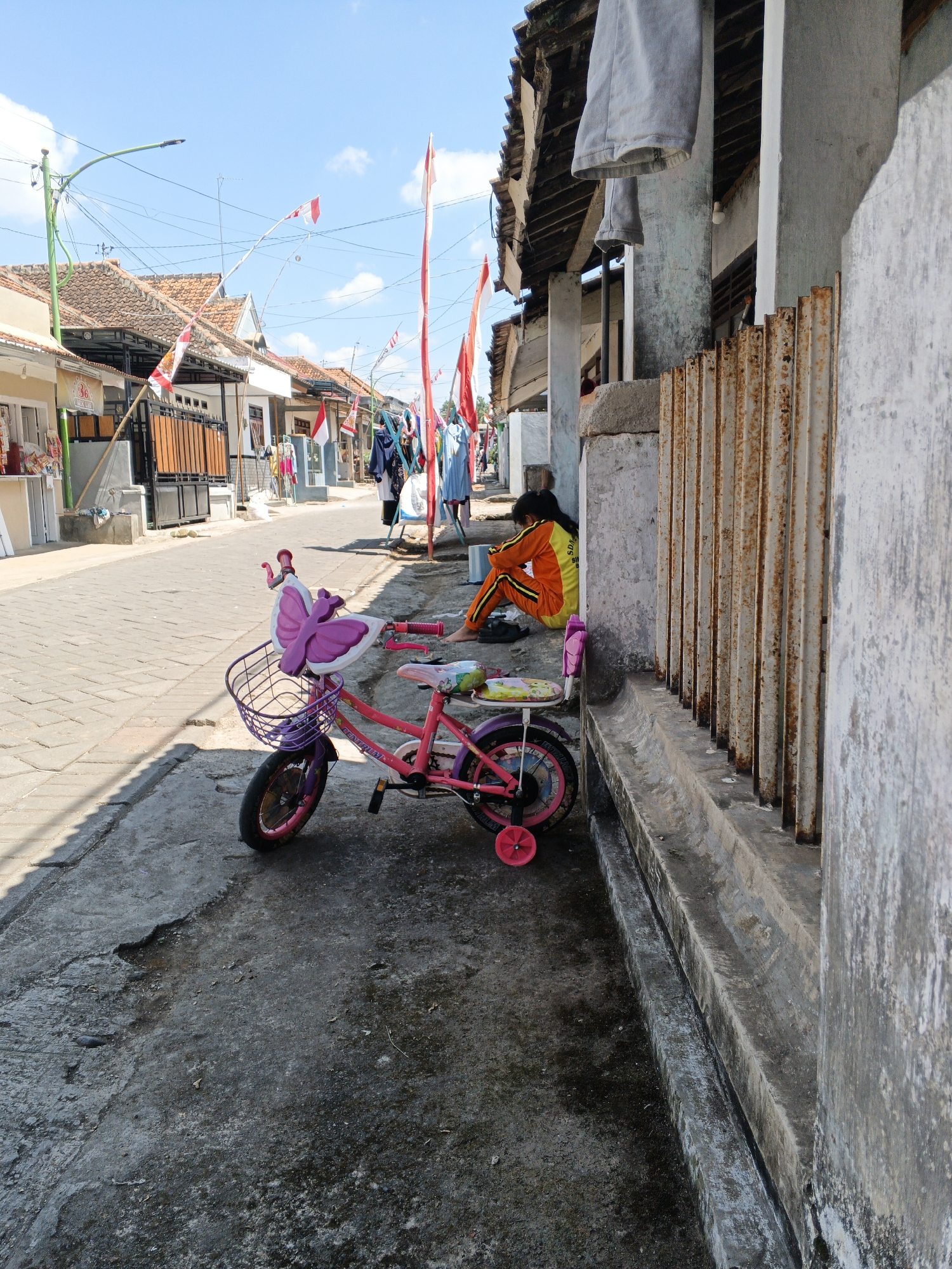 In Singolatren, Indonesia, a child sits on a curb in front of a weathered, rusty metal fence. The child, dressed in an orange and yellow tracksuit, appears to be looking at something in their lap. To the left of the child, a pink children's bicycle with purple accents and a butterfly decoration is parked on a paved street. Red and white Indonesian flags are visible along the street, strung between poles and attached to buildings, suggesting a celebration or holiday. The street is lined with buildings, some with tiled roofs and open verandas. Clothes are hung out to dry from an upper story, indicating a residential area. The sky is blue with scattered clouds, and the sun casts distinct shadows on the ground, indicating it is daytime.