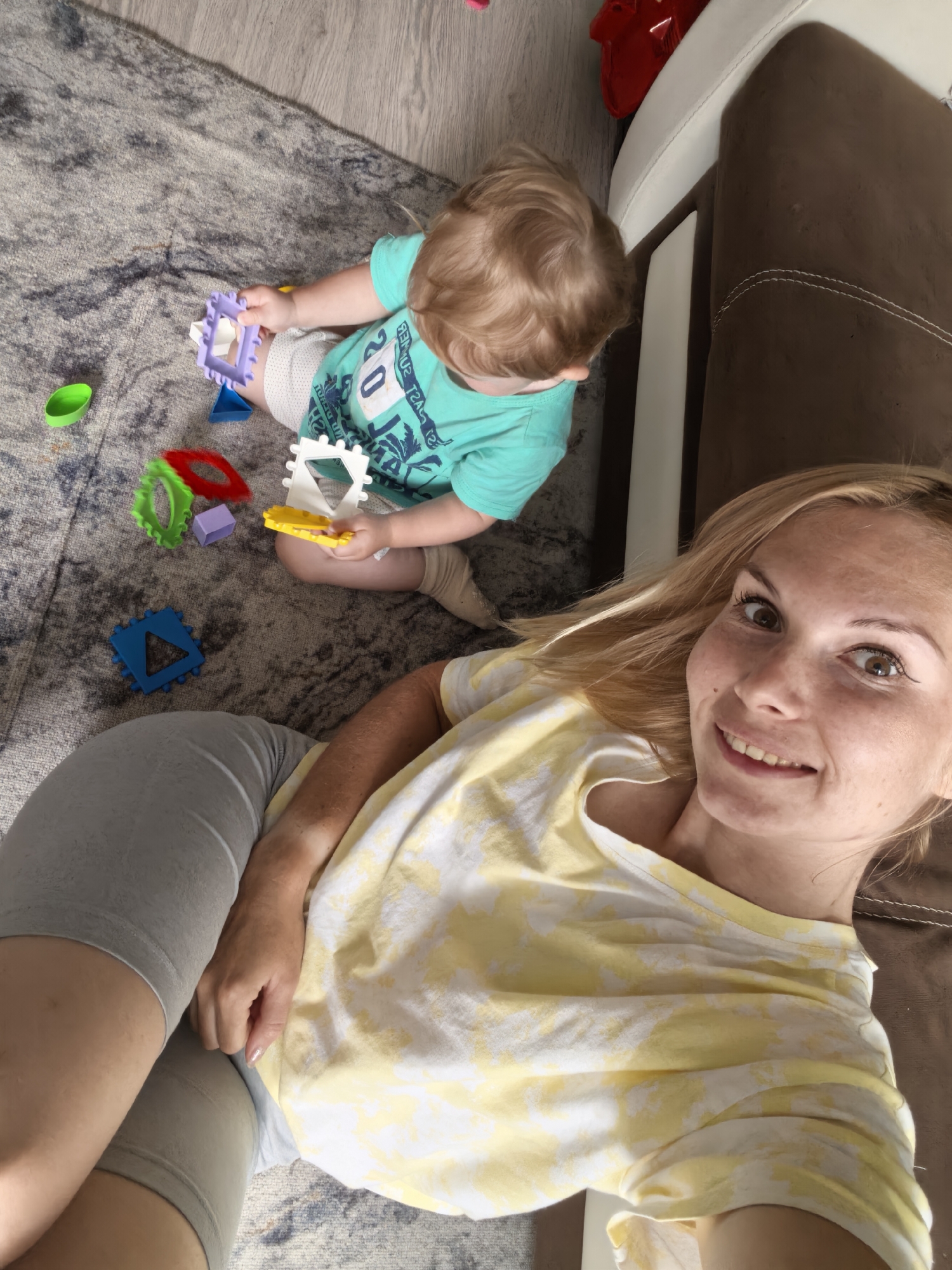 A high-angle medium shot captures a mother and child playing on a rug in front of a brown and white sofa. The child, in a turquoise t-shirt and white shorts, is on the floor, surrounded by colorful building blocks. The mother, in a yellow and white tie-dye shirt and grey shorts, is lying next to the child, looking at the camera. The scene appears to be in a living room. The overall impression is one of domesticity and playtime.