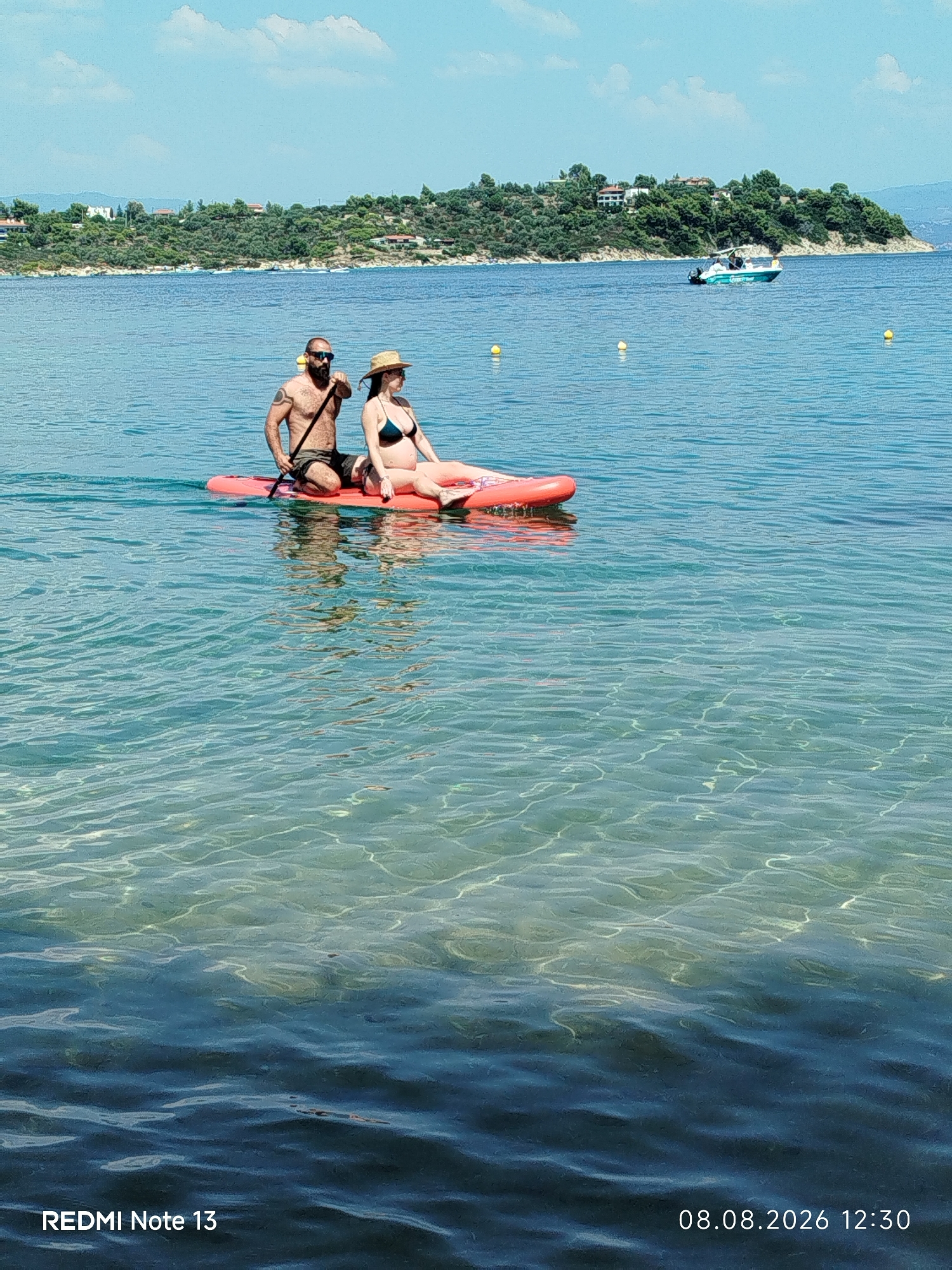 In Sithonia Municipal Unit, Greece, two people are on a paddleboard in clear blue water. A man with a beard and sunglasses is kneeling at the back of the board, holding a paddle. A woman wearing a straw hat and a dark bikini is sitting in front of him, resting her arm on her leg. The paddleboard is pink and floating on the water. In the background, a green, hilly coastline with trees and houses can be seen. A small white boat is visible on the water near the shore. The sky is blue with a few white clouds. The date and time stamp at the bottom right of the image reads "08.08.2026 12:30". The text "REDMI Note 13" is also visible at the bottom left.