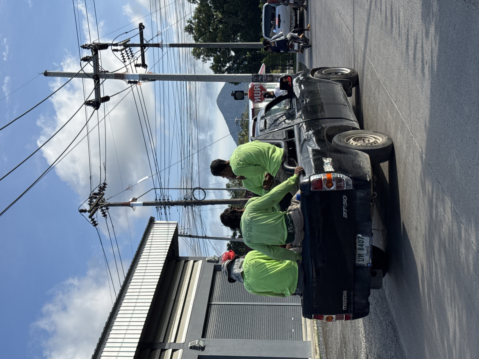 In the image, a black Mazda BT-50 pickup truck is driving on a road in ตำบลเกาะพะงัน, Thailand. Four individuals, likely workers, are seated in the bed of the truck. Three of them are wearing bright green long-sleeved shirts with "ECOLSF FACTORY" printed on the back. The truck is in motion, indicated by the blurred road surface and the shadows cast by the vehicle and other objects. The environment includes a building with a corrugated metal facade on the left, utility poles with numerous wires overhead, and lush green trees in the background. A mountain is visible in the distance. Traffic signals and other vehicles, including motorcycles and cars, are present on the road, suggesting a moderately busy street. The sky is blue with scattered white clouds. The license plate of the truck is visible and reads "บพ 8407".